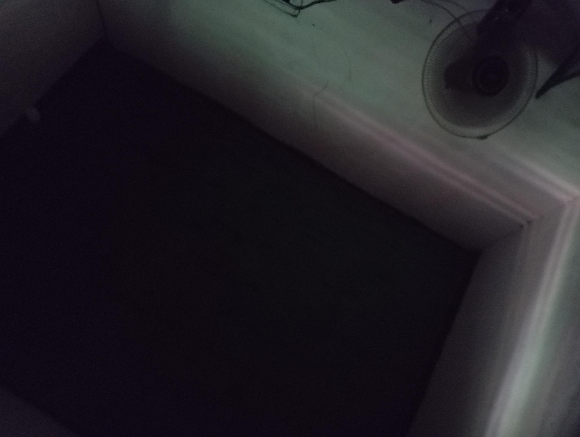 In Rejosari, Indonesia, this image shows an indoor scene with poor lighting. The upper portion of the frame is dominated by a dark, likely ceiling area. In the lower left, a partially visible electric fan is present. The walls appear to be a light color, possibly white or pale green, with some subtle light reflections suggesting a light source from the left. The overall impression is of a dimly lit room.
