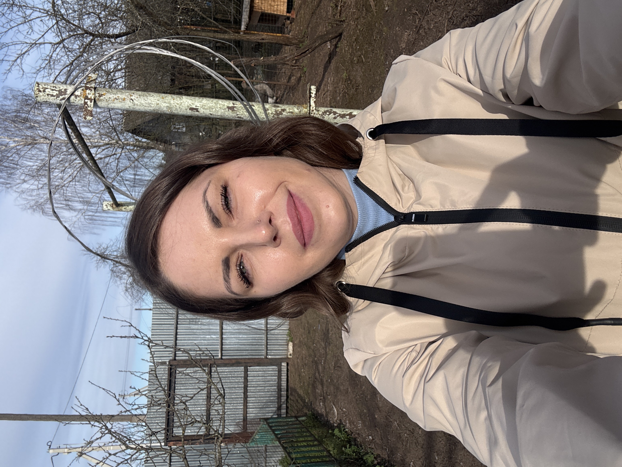A woman stands outdoors in Razdolye, Russia. She is wearing a light-beige jacket with black accents and a blue top underneath. A corrugated metal fence and bare trees are visible in the background, along with structures. The ground appears to be covered with dirt and vegetation. The sky is overcast.