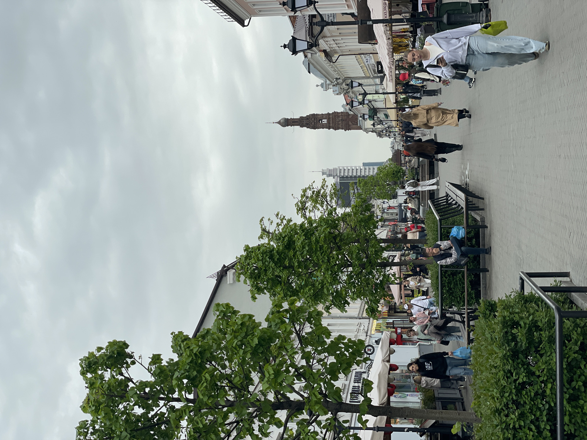 The image shows a street scene in Kazan, Russia, oriented with the tower aligned centrally. People are walking or sitting on benches along a wide pedestrian area. The sky is overcast. Buildings line both sides of the street. Trees and bushes provide greenery along the pedestrian area. A tall, slender tower is visible in the distance.