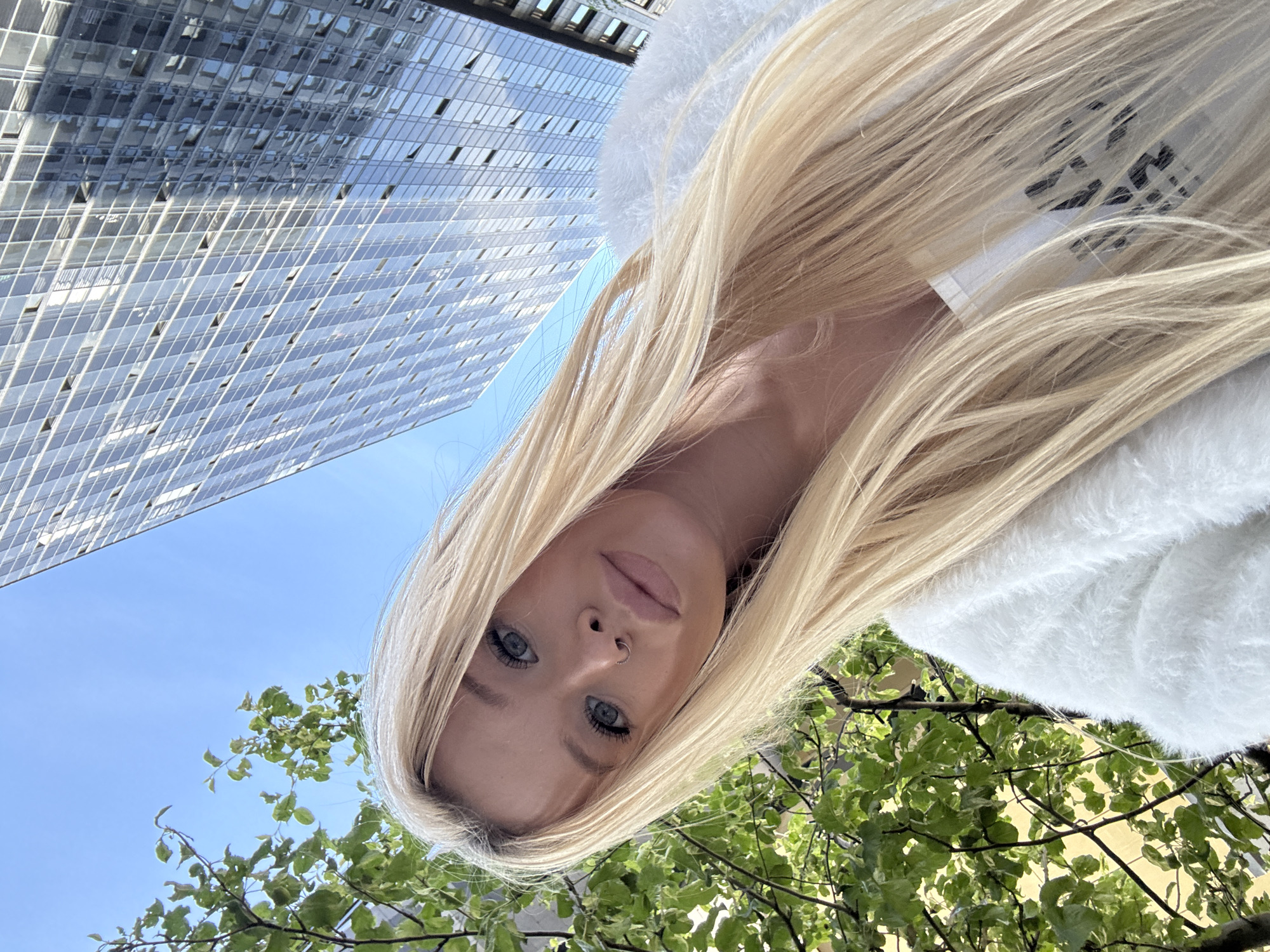 A young woman with long blonde hair is taking a selfie outdoors. She is wearing a white fluffy garment, possibly a jacket or scarf, and her face is turned towards the camera. She has blue eyes, light-colored eyebrows, and a small nose ring. Her lips are slightly parted, and she appears to be looking directly at the lens.
The background of the image features a clear blue sky, the reflective glass facade of a modern skyscraper, and the lush green foliage of trees. The skyscraper's windows create a geometric pattern that contrasts with the organic shapes of the leaves. The composition is angled, giving a sense of looking up from ground level.
The lighting suggests it is daytime, likely sunny, given the brightness and the clear blue sky. The presence of a modern building and trees points to an urban setting.
There is no visible text in the image.