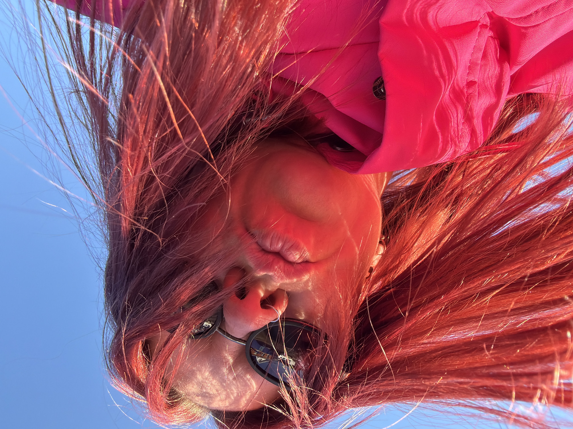 The image is an upside-down close-up of a person with red hair wearing sunglasses and a pink top. The person has pursed lips. The background is a clear blue sky. The reflection in the sunglasses suggests an urban environment with buildings. While it's impossible to definitively pinpoint the location as Chaikovsky, Russia, based on the visual information alone, the reflection in the sunglasses could potentially contain contextual clues for a more precise determination if analyzed further.