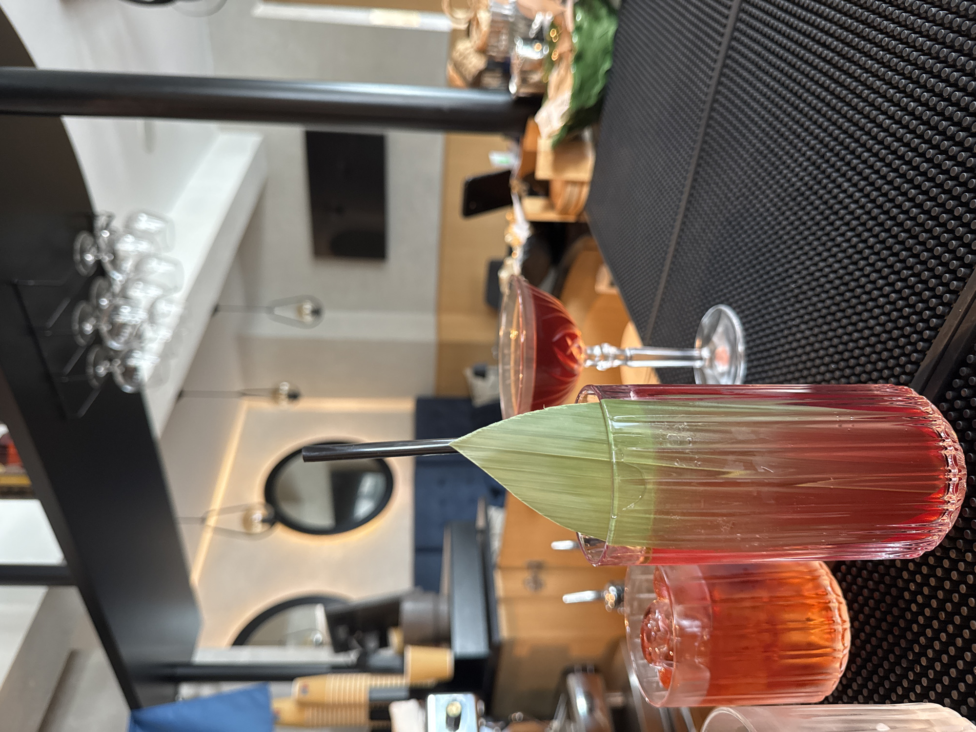 In a dimly lit bar, likely in Boreti, Montenegro, two vibrant red cocktails are prominently displayed on a black textured bar counter. The cocktail on the right, in a tall fluted glass, is garnished with a large, vibrant green leaf and a dark straw. To its left, a shorter, wider glass holds another red drink with ice. In the background, out of focus, a sleek espresso machine sits on a wooden counter, adjacent to a blue cushioned bench. The bar's ambiance is enhanced by subtle lighting, with glowing pendant lamps and circular mirrors adorning the wall. A television screen is visible in the distance, adding to the modern yet relaxed setting. The overall scene suggests a sophisticated establishment, potentially a hotel bar or a high-end lounge, offering a tranquil escape.