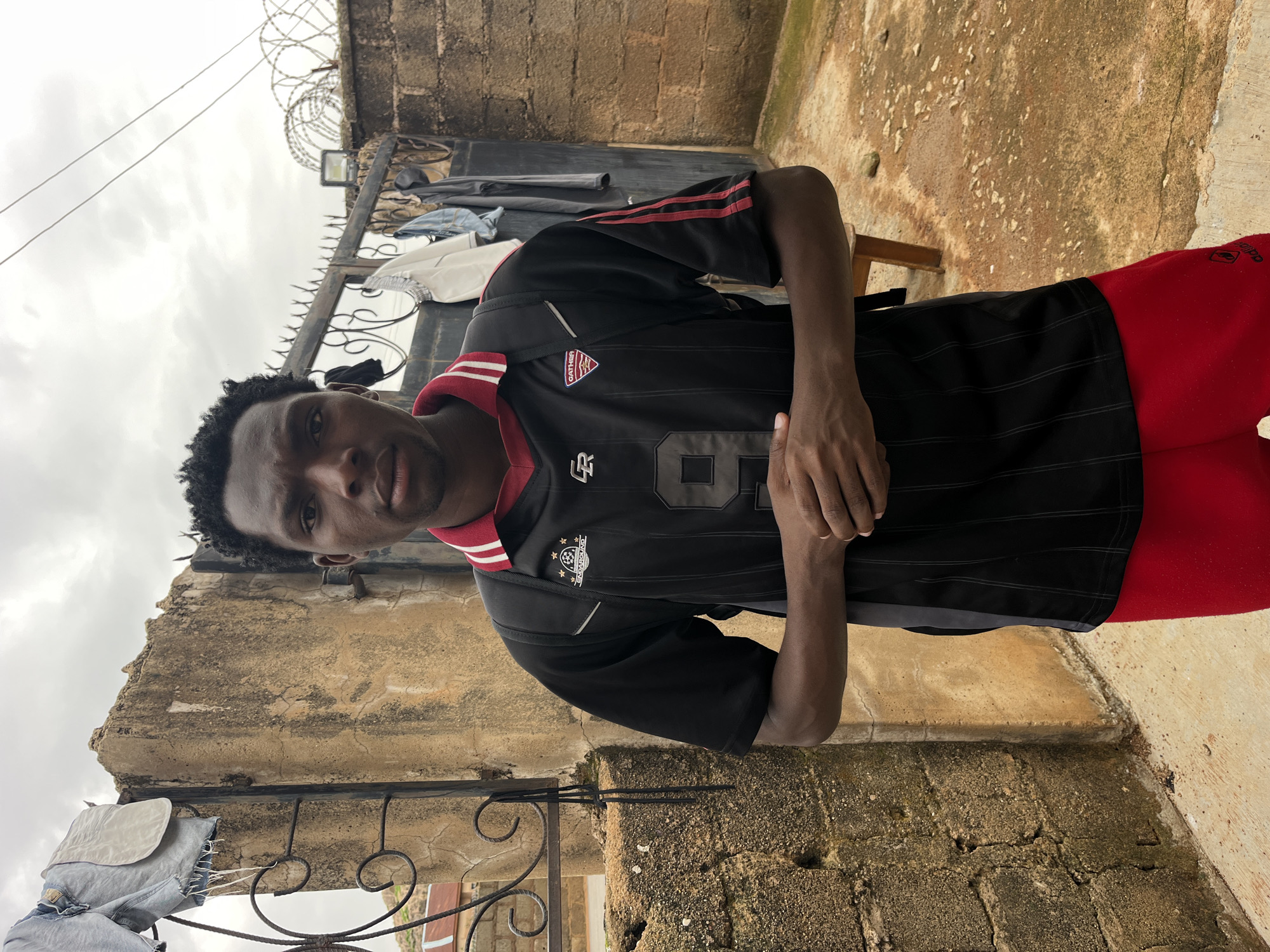 A young Black man stands in front of a weathered concrete wall and a metal gate. He is wearing a black t-shirt with red trim and red sweatpants. He has a black backpack on. Clothes are hanging on the gate. The ground is concrete. The sky is overcast.