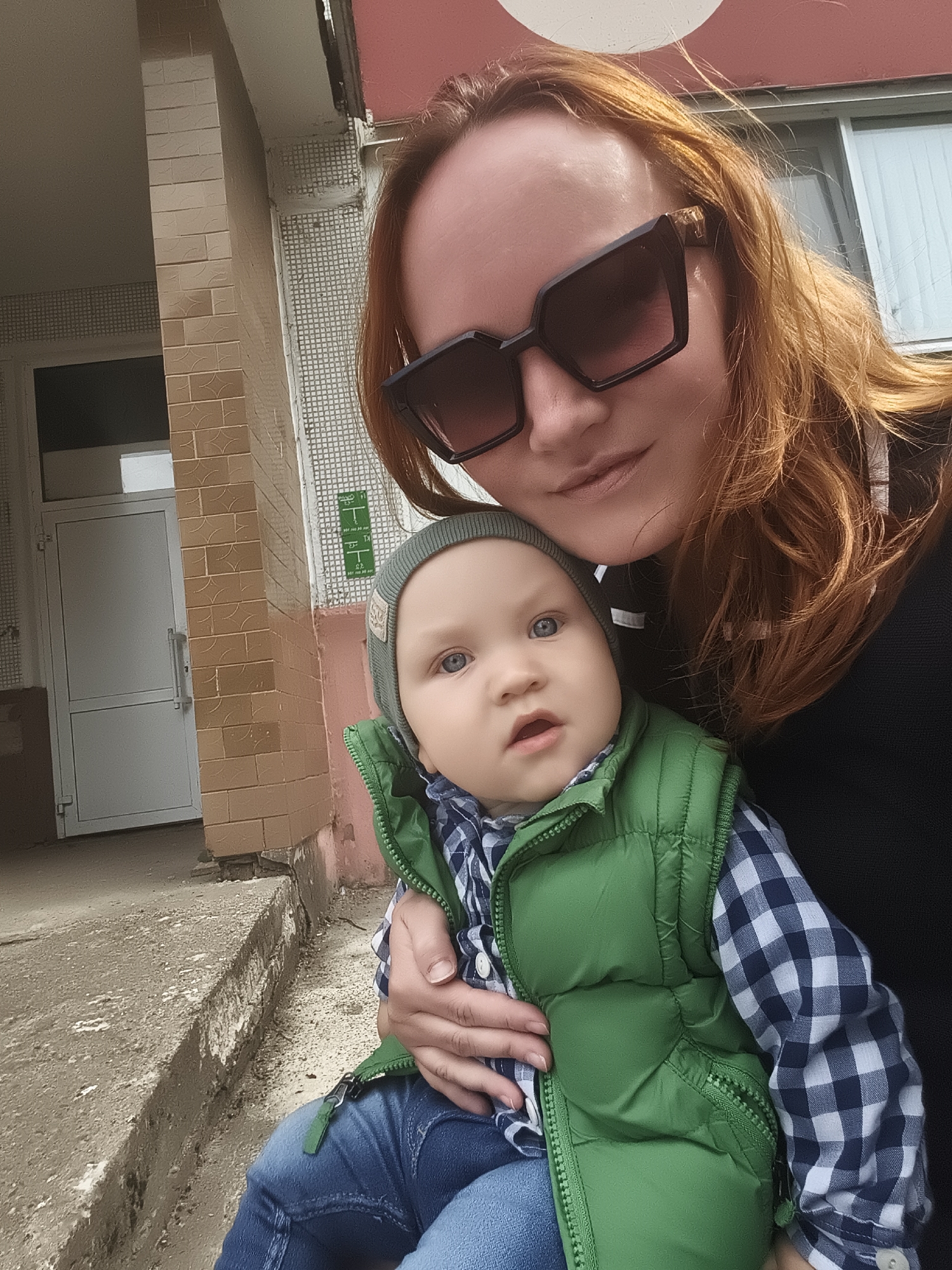 A woman with red hair, wearing large black sunglasses, is taking a selfie outdoors with a baby. The baby, wearing a grey hat, a green vest, and a blue and white checkered shirt, is held in the woman's arms. The background shows the exterior of a brick building with a white door. The location is identified as Chaikovsky, Russia. The scene appears to be a casual, everyday moment.