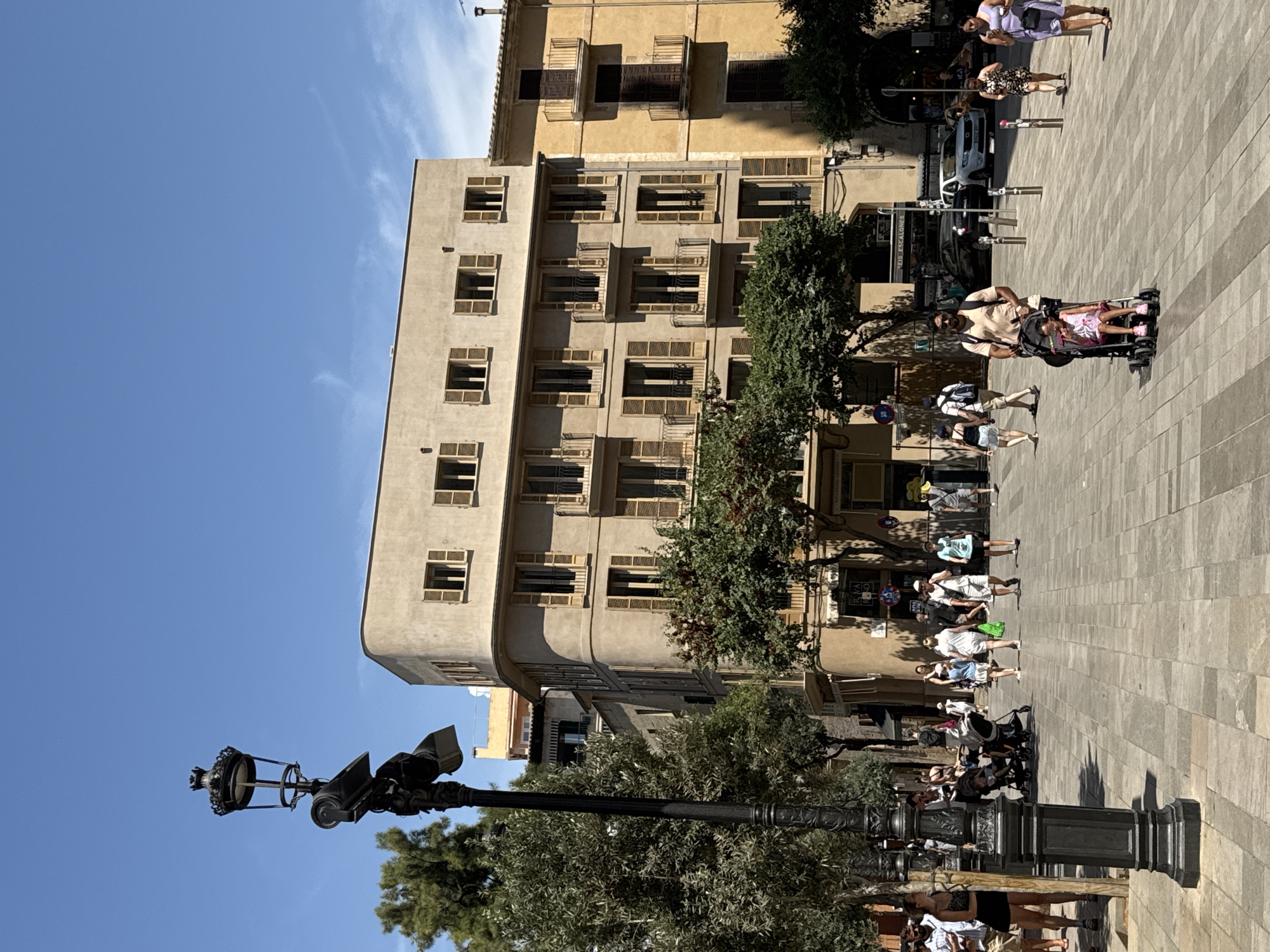 The image depicts a sunny day on a wide, pedestrianized street in Palma de Mallorca, Spain, likely in a commercial or historical district. The street is paved with light-colored square stones, and a tall, ornate black lamppost with a surveillance camera attached stands prominently in the left foreground.
Numerous people are walking along the street in both directions. In the foreground, a man is pushing a stroller with a young child in it. Further down the street, groups of individuals, some appearing to be tourists, are strolling, some with shopping bags. Many are dressed in light, summer clothing.
The background is dominated by several buildings. A large, light-beige, modern-style building with rounded corners and multiple floors featuring numerous windows with brown shutters occupies the central part of the frame. To its right, a narrower, taller building with a yellow facade and balconies with dark railings is visible. Trees with green foliage are interspersed along the street and in front of the buildings, providing some shade. The sky is clear blue with a few wispy clouds. Cars are visible in the background on a road beyond the pedestrian area. Signs are present on the buildings, suggesting commercial establishments.