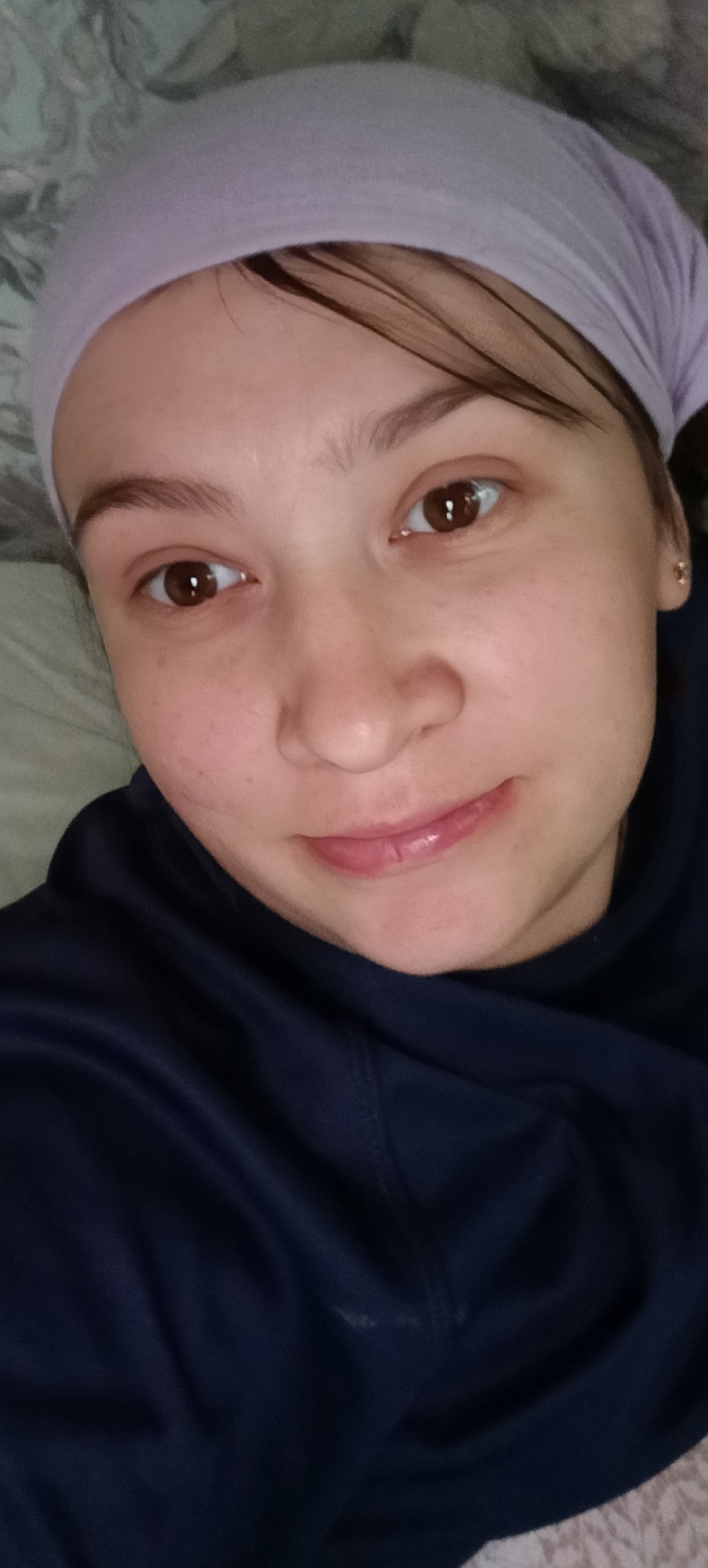 A close-up, overhead shot shows a young woman lying down, looking upwards. She is wearing a light purple headscarf and a dark blue hooded sweatshirt. Her brown eyes are wide and looking directly at the camera. She has light skin with some freckles and light pink lips that are slightly parted. The background consists of a patterned textile, likely bedding. The image quality is moderate, with some graininess and soft focus. No information is available regarding the location in the city of Khokhly, Russia.