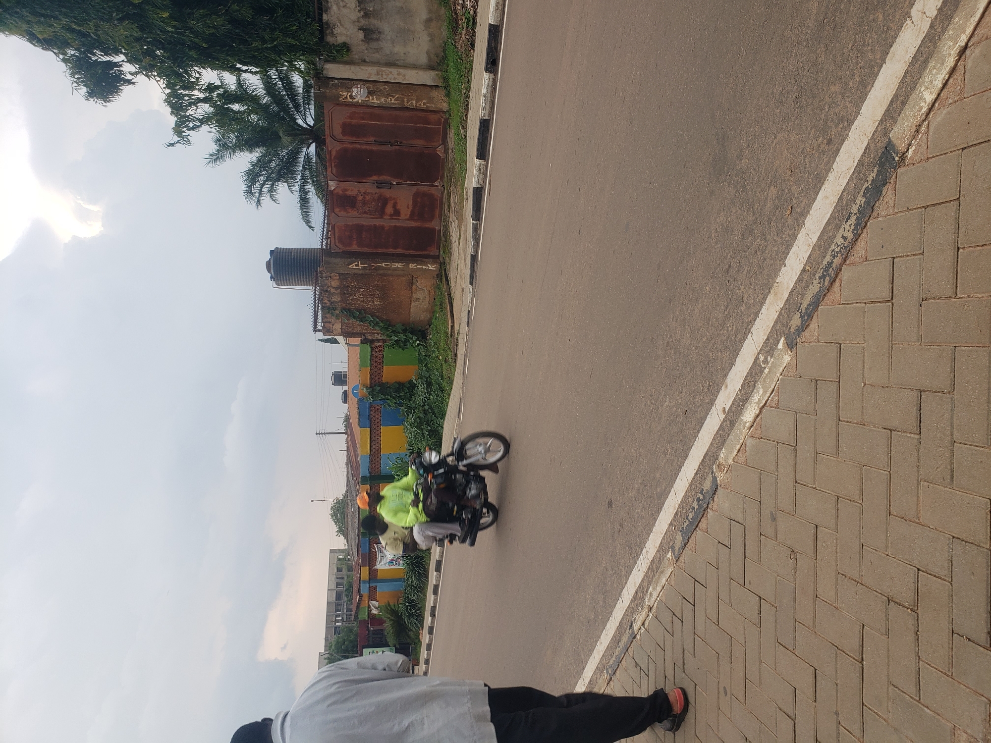 In Narayi, Nigeria, a motorcyclist wearing a bright green hoodie and an orange cap, with a passenger behind him, is traveling on an asphalt road. The motorcyclist is blurred due to motion. To the left of the road, a pedestrian in a light-colored shirt and dark pants is partially visible, facing away from the camera. The road has a white line marking its edge. On the right side of the road, there is a weathered brick wall with large, rusty metal gates. Behind the wall, a water tank and palm trees are visible. The sky is overcast with diffused light.