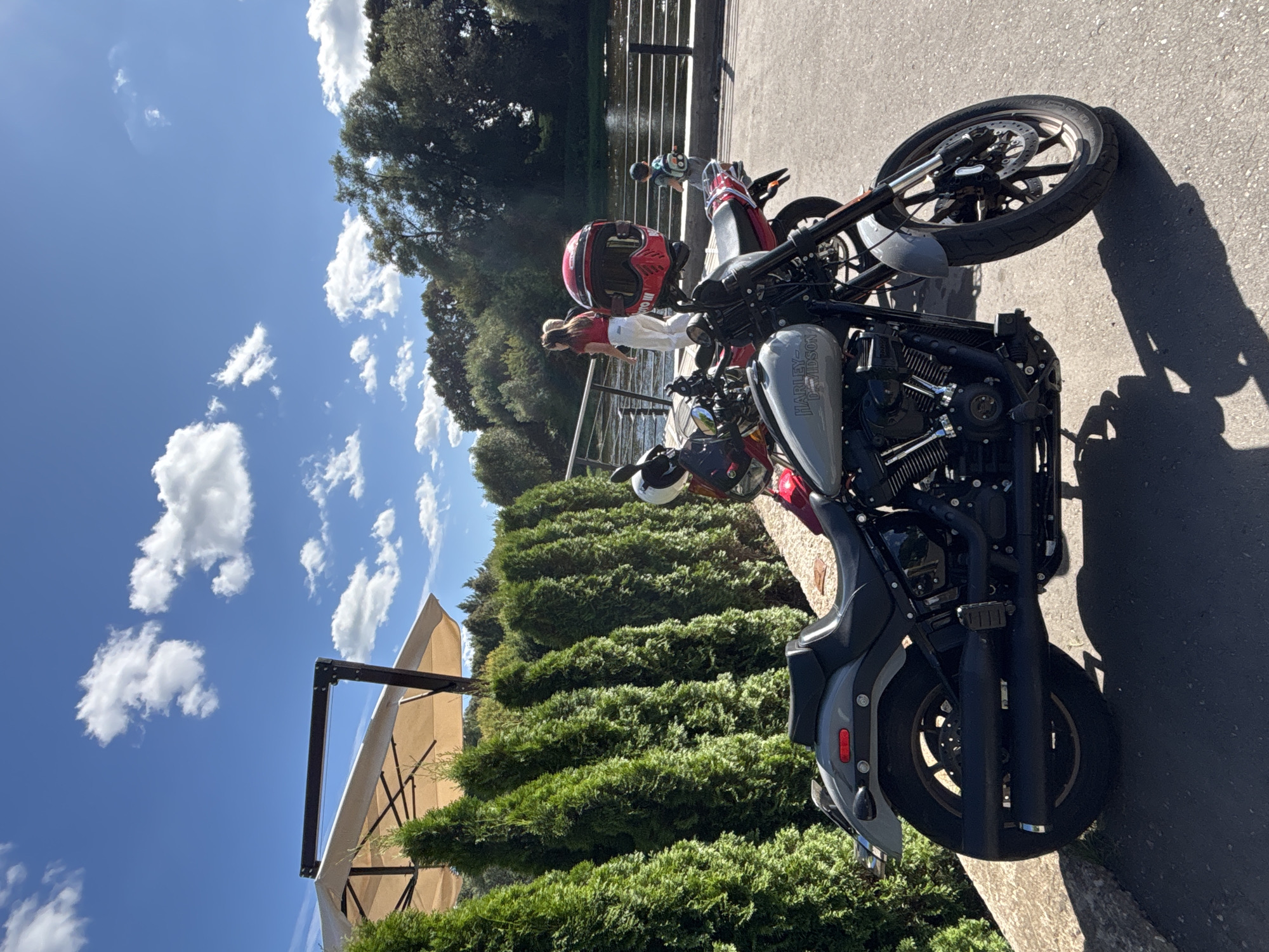A grey Harley-Davidson motorcycle is parked on asphalt in front of a row of trimmed green trees. In the background, a river or body of water is visible with a pedestrian walkway along its edge. Several people are present on the walkway, including two women and a child. A red motorcycle helmet is attached to the handlebars of the grey motorcycle. The sky is blue with scattered white clouds. The scene appears to be a public outdoor space, potentially a park or waterfront area in Zvenigorod, Russia.