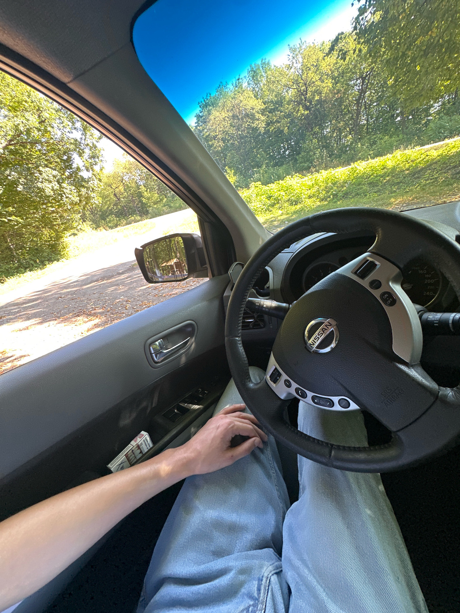 An interior view from the driver's seat of a Nissan vehicle shows a person's arm resting on the open door and light blue jeans. The steering wheel, dashboard with a Nissan logo, and a pack of cigarettes in the door pocket are visible. Outside the car, a dirt road winds through a sun-drenched forest with green trees. The sky is a clear, bright blue. The location is the city of Shatava, Ukraine.