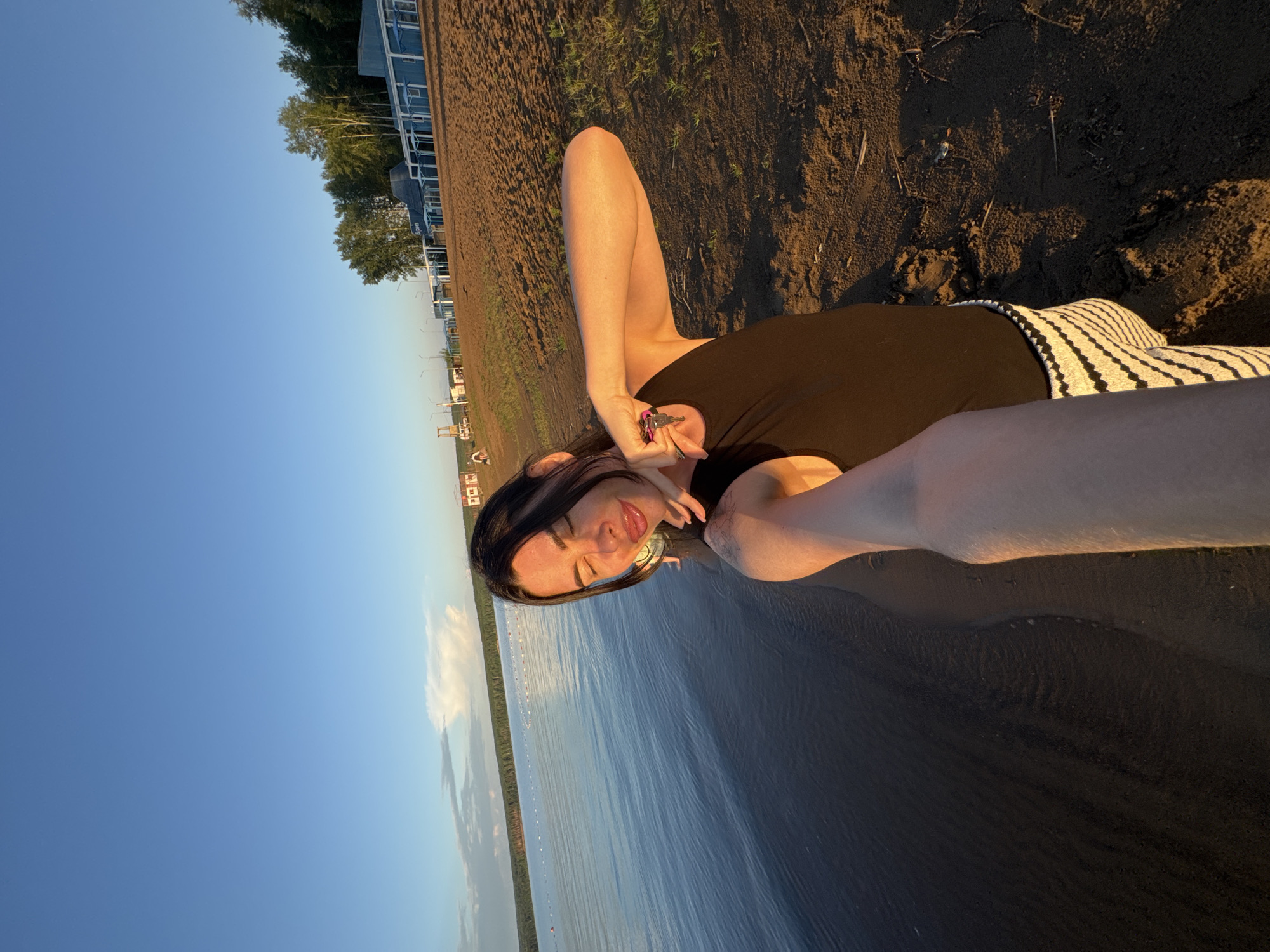 A selfie is taken by a woman with dark hair on a sandy beach next to a body of water. She is wearing a black top and striped pants. Her eyes are closed and she is smiling, holding keys in her hand. The beach appears to be in Chaikovsky, Russia, with a line of buoys visible in the water and a blue building with a sign in Cyrillic script in the background, along with trees and a clear blue sky. The lighting suggests it is late afternoon or early evening.