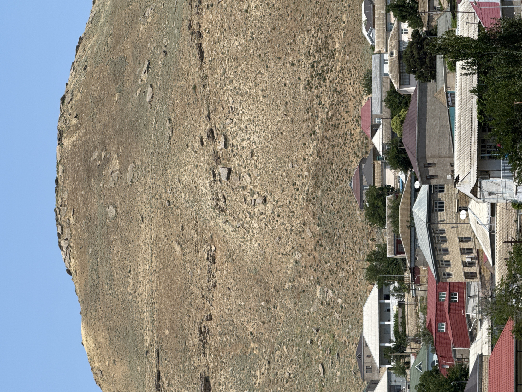 In Lokbatan, Azerbaijan, a semi-arid hillside settlement is depicted under a clear blue sky. The scene is dominated by a large, barren mountain that slopes down towards a cluster of houses. The mountain's terrain is a mix of dry grass, soil, and scattered rocks. The houses are varied in size and design, with many featuring flat or sloped roofs in shades of gray, white, and red. Some houses are constructed with stone or concrete, while others appear to be painted in lighter colors. Trees and greenery are interspersed among the dwellings, suggesting pockets of vegetation in the otherwise dry environment. The image captures a view of residential buildings nestled against the base of the imposing natural landscape.