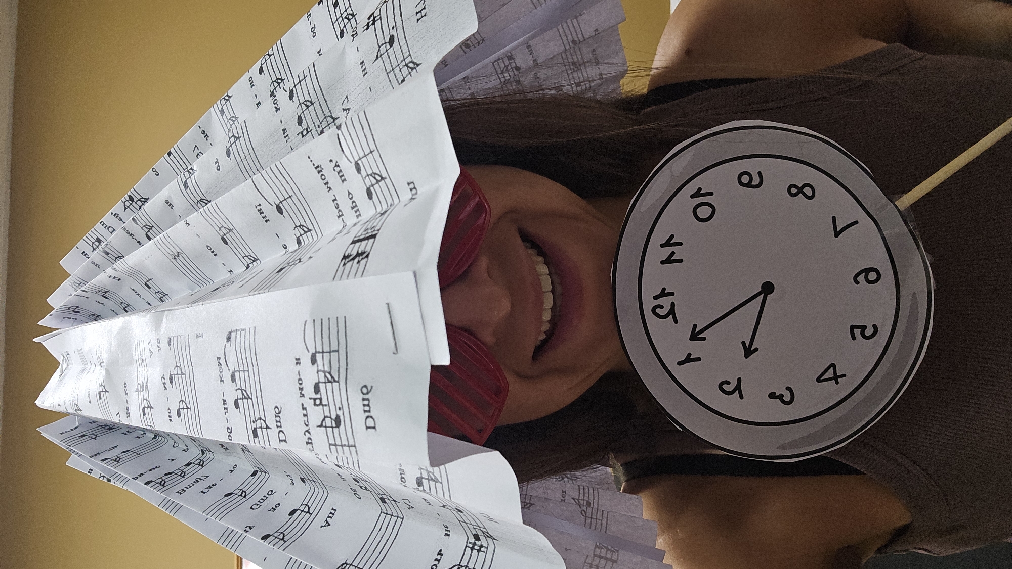 A person is wearing a hat made of folded sheet music and red novelty glasses. They are holding a clock with Roman numerals on a stick in front of them. The background is a plain yellow wall. The image appears to be taken indoors.