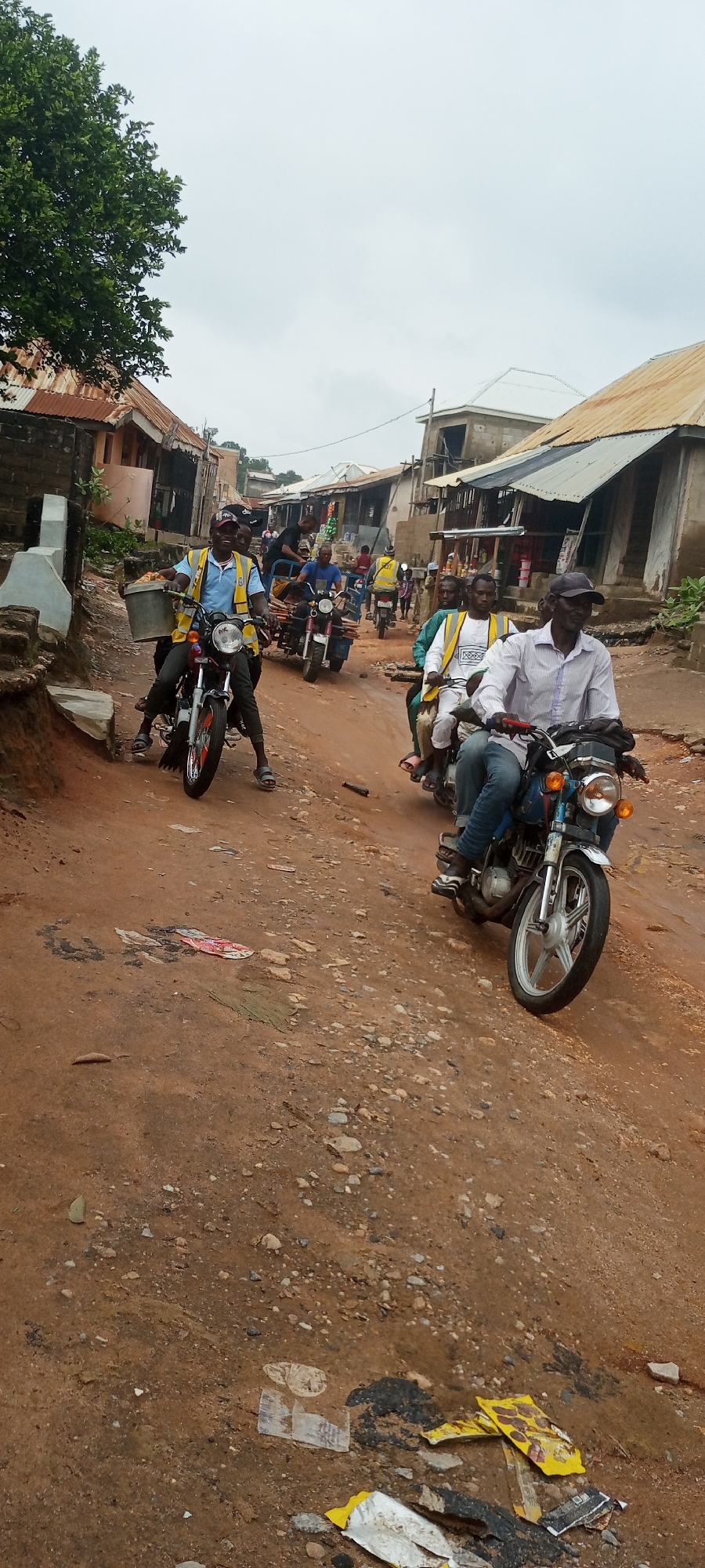 This is a ground-level, vertical, medium-shot photograph taken on a dirt road in Zango-ndaji, Nigeria. The scene depicts a procession of motorcycles carrying multiple passengers through a village setting. The road is a mixture of reddish-brown earth and scattered stones, with some patches of mud indicating recent rain. Buildings line both sides of the road, characterized by corrugated metal roofs and walls made of various materials, suggesting a developing or rural area.
In the foreground, a man wearing a light blue short-sleeved shirt and a bright yellow reflective vest is riding a red motorcycle. He is looking forward and smiling, with a large gray bucket attached to the front of his motorcycle. He appears to be the leader of the procession. Behind him, several other motorcycles are visible, also carrying passengers. Some of the passengers are wearing similar yellow reflective vests, suggesting they might be part of a group or working in a similar capacity. One motorcycle in the mid-ground has a wooden structure attached to its rear, possibly for carrying goods.
The sky is overcast, contributing to a muted lighting condition. The environment appears to be a typical village street with a mix of residential and commercial structures. Litter, including plastic wrappers and debris, is scattered along the roadside, a common sight in many urban and semi-urban areas. The overall impression is one of community movement and daily life in a Nigerian village.