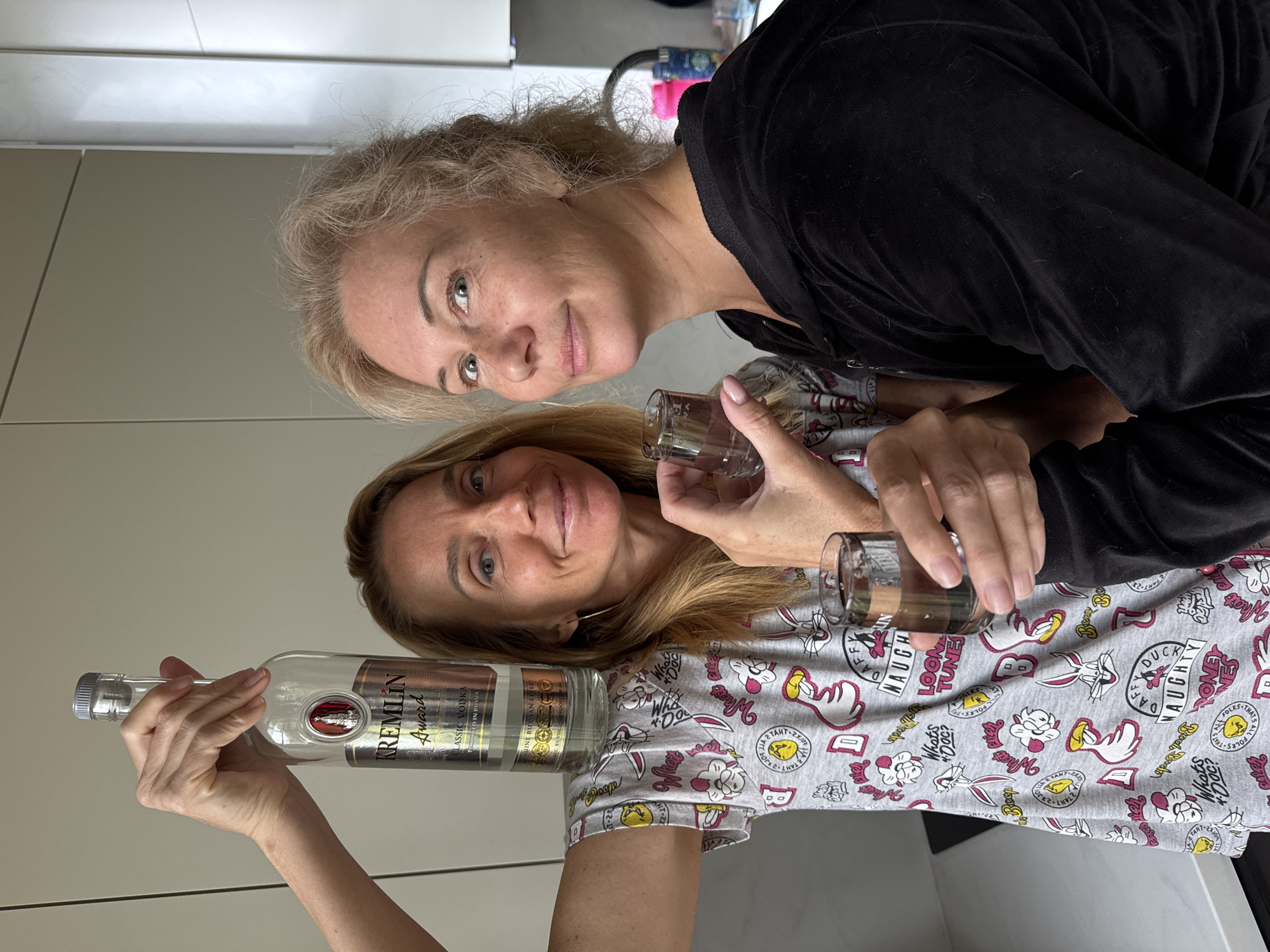 Two fair-skinned women are posing indoors, one holding a bottle of "Kremlin Award Classic Vodka Produced in Russia" and two shot glasses. The woman holding the bottle is in the foreground, wearing a grey pajama top patterned with cartoon characters. The second woman, positioned behind, wears a black top. The location is potentially the city of Steklyannyi, Russia, based on the origin of the vodka brand. The background is a light-colored wall or cabinet.