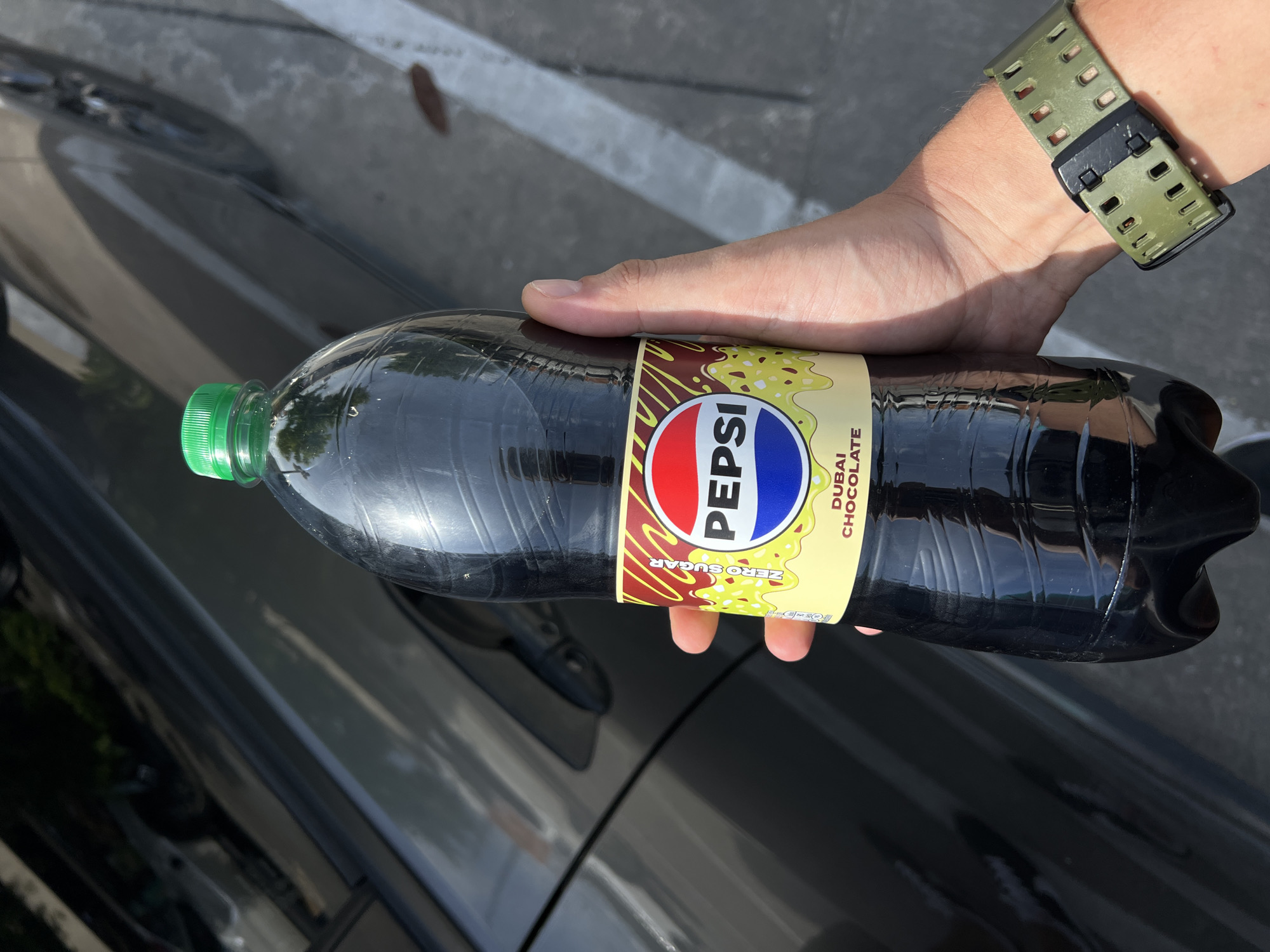 A person holds a bottle of Pepsi Zero Sugar Dubai Chocolate flavor in front of a dark-colored car. The bottle has a green cap and a yellow label with the Pepsi logo and text indicating "Zero Sugar" and "Dubai Chocolate". The person is wearing a green watch on their left wrist. The background shows the side of a car and a paved surface with a white line.