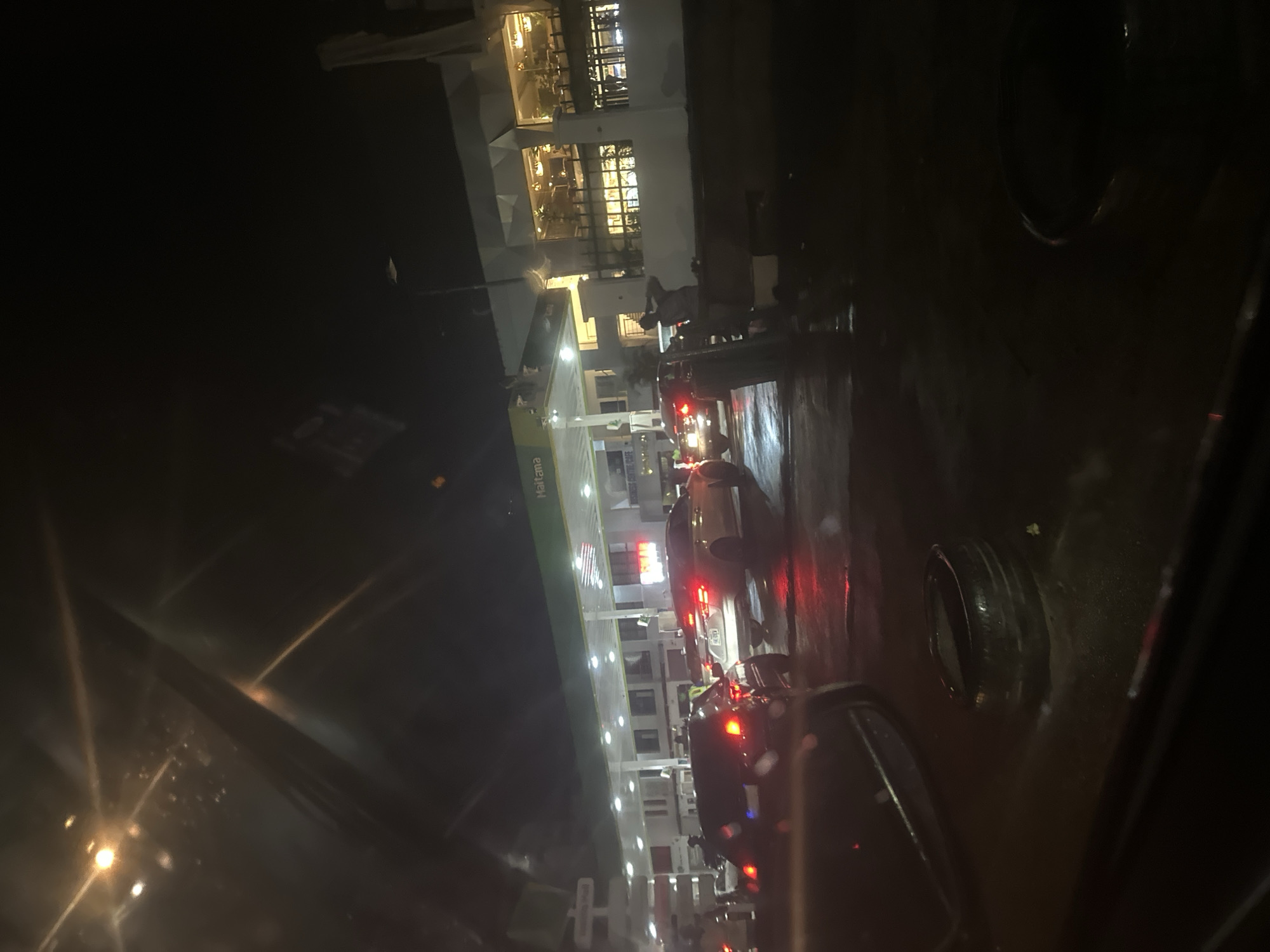 This image depicts a nighttime scene at a gas station in Abuja, Nigeria. The gas station appears to be branded with "Haitama" visible on the canopy. Several cars are lined up, suggesting a queue, possibly for fuel. The lighting from the gas station and car headlights creates reflections on the wet pavement, indicating recent rain or a damp environment. In the background, a building with illuminated windows is visible, suggesting it might be a restaurant or shop associated with the gas station. One individual is standing and appears to be adjusting their clothing, while others are inside or near vehicles. The overall atmosphere is that of a busy, functional urban area at night.