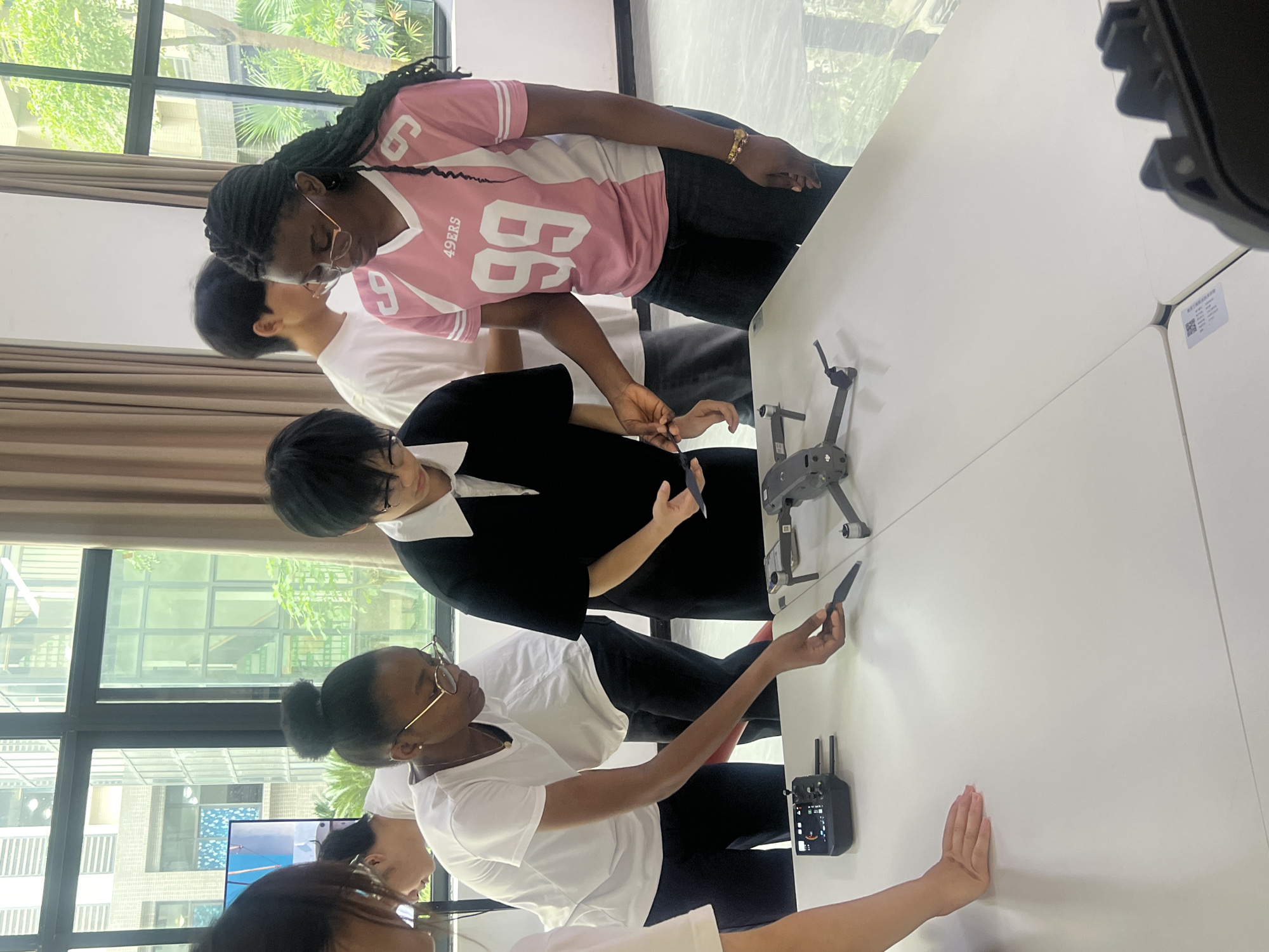 A group of people are gathered around a white table, inspecting a drone. One person is holding a drone propeller. The people are standing indoors near a window with a view of trees and buildings. The location is Jiangjin, China.