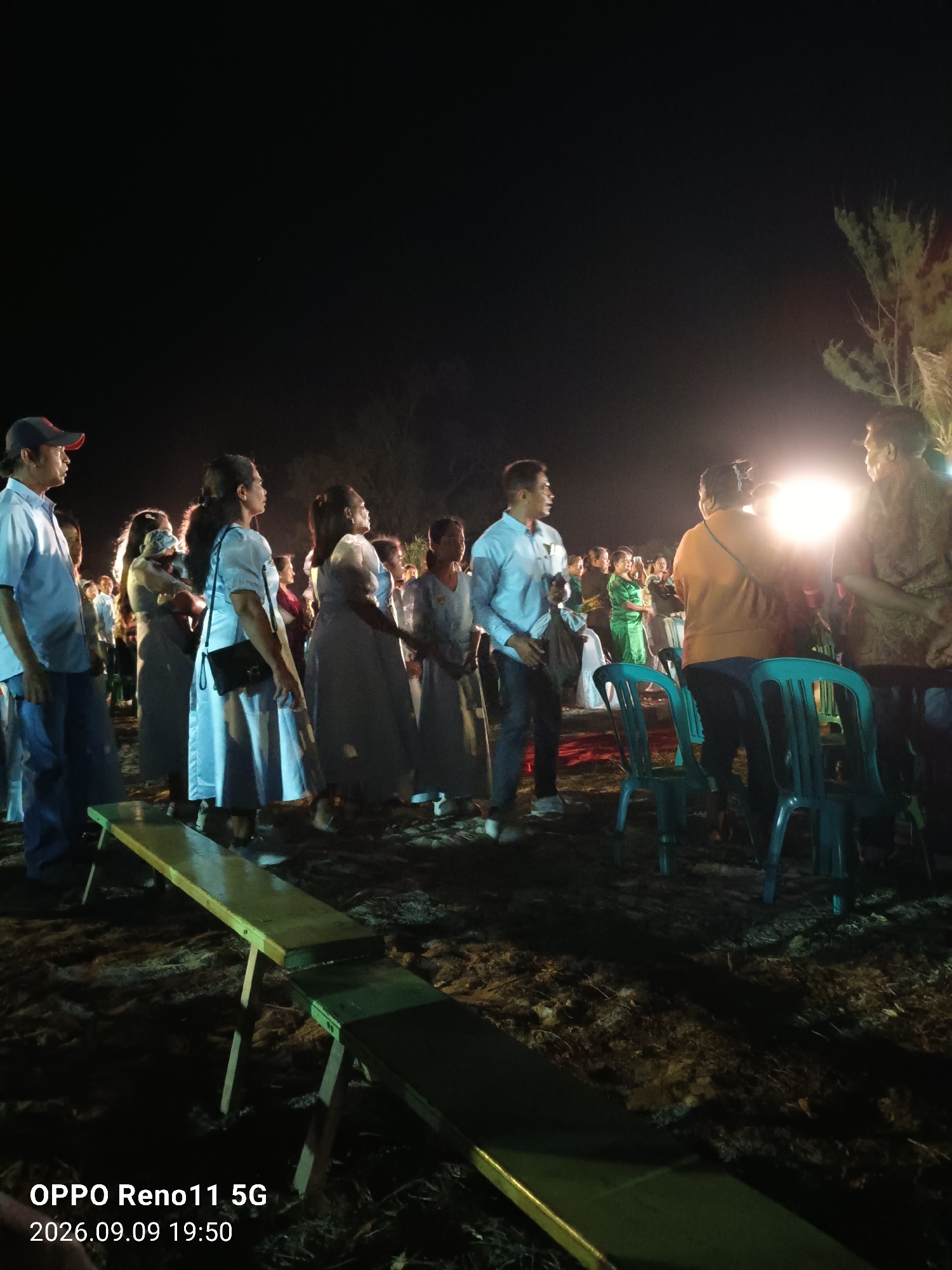 The image captures an outdoor gathering at night, likely in the city of Otan, Indonesia. The scene is illuminated by a bright light source, possibly a spotlight or a powerful lamp, situated in the mid-ground to the right. Several individuals are visible, with a group of women in light-colored dresses standing in the center and to the left. One woman in the foreground is wearing a light blue dress and carrying a black purse. To the far left, a man in a blue shirt and a baseball cap is looking to his right. On the right side, two men are standing, one in a patterned shirt and the other in a light blue shirt. In front of the gathering, a wooden bench with a yellow surface and green legs is placed on the ground, which appears to be sandy or earthy. Several plastic chairs, primarily turquoise, are scattered around the area. The background is dark, with some sparse vegetation visible on the right. The timestamp in the bottom left corner indicates the image was taken on September 9, 2026, at 19:50.