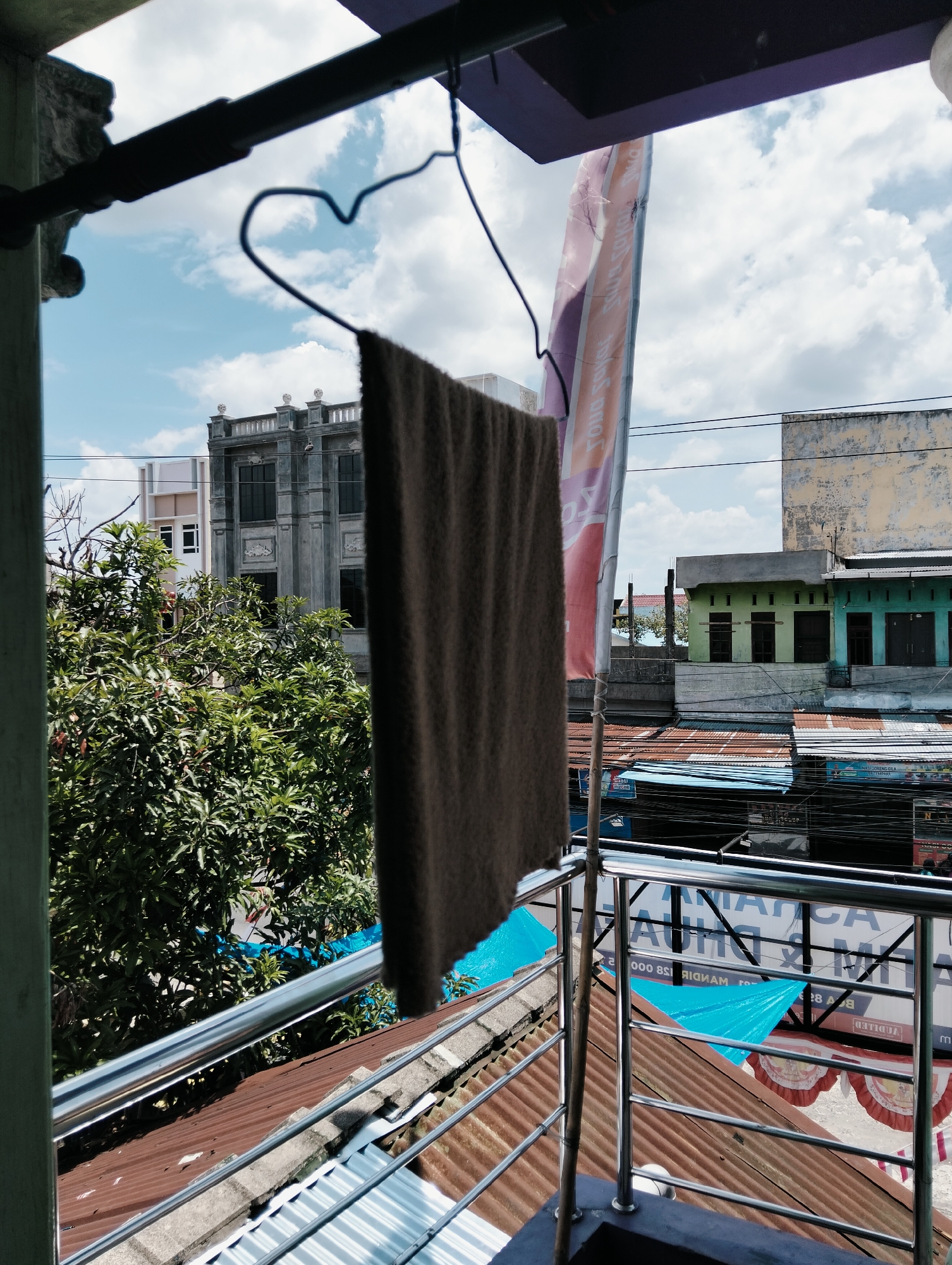 This is an outdoor, eye-level shot of a balcony railing in the City of Medan, Indonesia. A brown towel hangs from a wire hanger on a clothesline, partially obscuring a tall, colorful banner that reads "JAYA SUKA JAYA." In the background, a dense green tree is visible, followed by several buildings. One building is a large, gray, multi-story structure with decorative elements and many windows. To its right, a lighter-colored building stands next to it. Further into the background, there are green and turquoise buildings with closed doors and windows, and a yellow building. A network of electrical wires crisscrosses the scene. The sky is blue with scattered white clouds. The foreground shows a corrugated metal roof and a shiny metal railing.