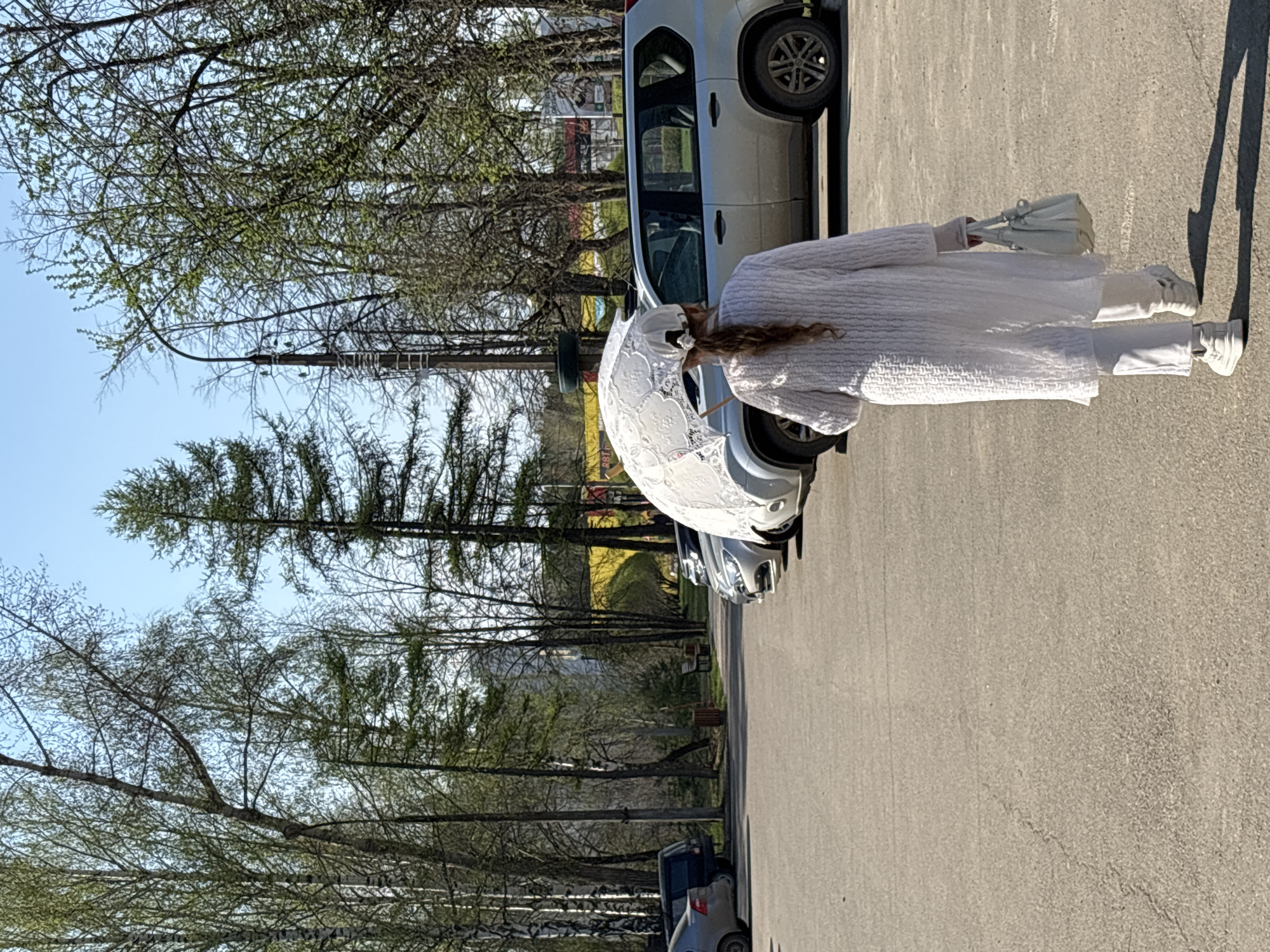 A woman in an all-white outfit, including shoes, pants, a textured top, and a lace parasol, stands on a gray paved surface. She also carries a matching white bag. The background consists of a row of parked cars, with silver and gray vehicles visible. Beyond the cars, there are trees with budding leaves, suggesting it is spring. The sky is clear and blue. The scene seems to be taken in a parking lot or a wide walkway near a road, as suggested by the cars parked alongside.
Based on location metadata associated with the image, the photo was taken in the city of Chaikovsky, Perm Krai, Russia.