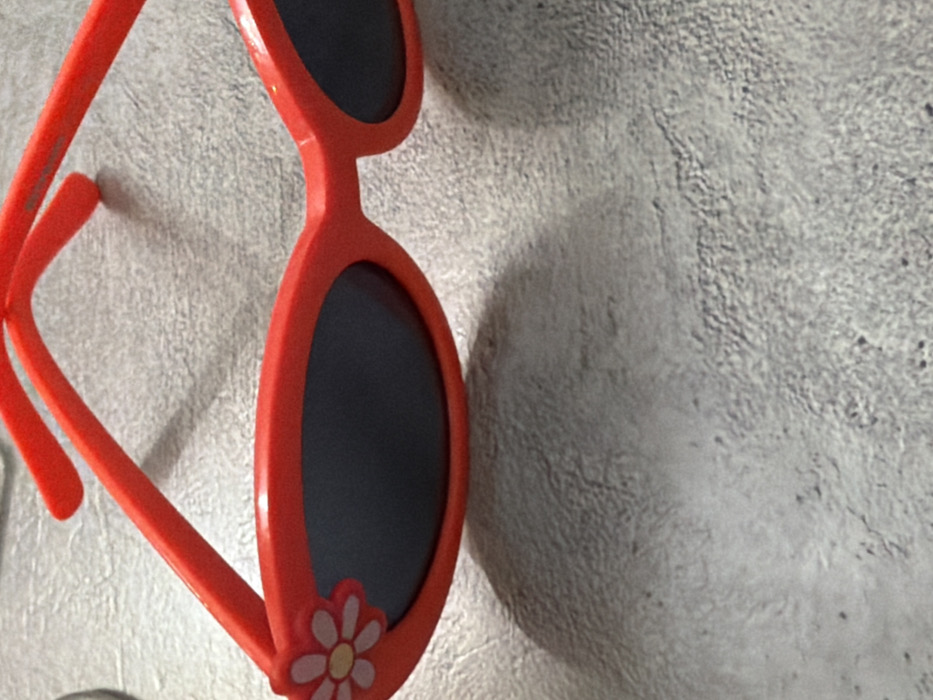 A pair of red-framed sunglasses rests on a textured, light grey surface in Komsomolskiy, Russia. The sunglasses feature dark, oval-shaped lenses. The left lens frame is adorned with a white and yellow daisy appliqué on its outer corner. The red temples extend upward and outward from the frame. Shadows from the sunglasses are cast onto the textured surface, which shows minor variations in tone and relief. No other objects or individuals are present in the scene.