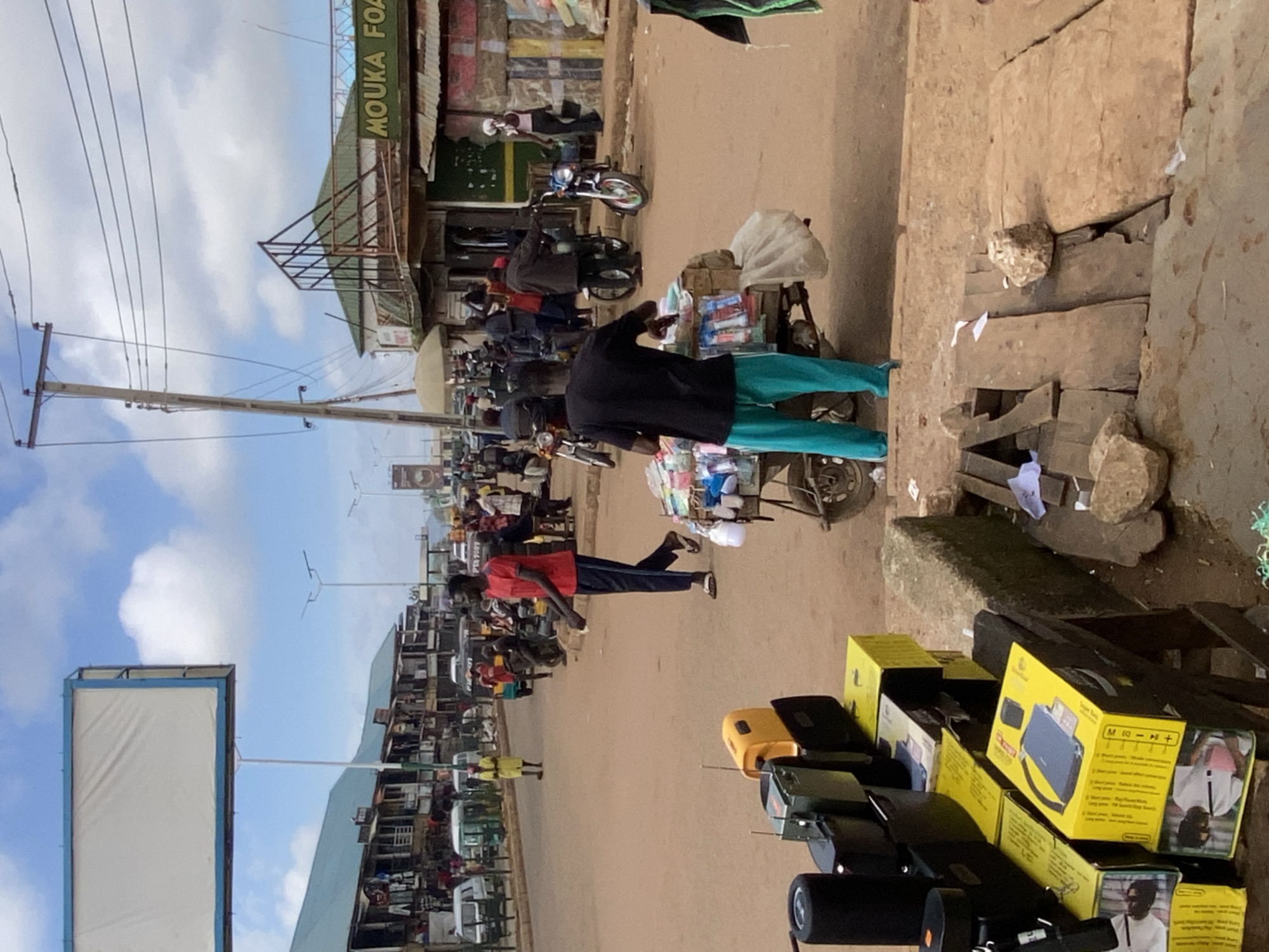 The image depicts a street scene in Bukuru, Nigeria, characterized by a bustling market environment under a partly cloudy sky. The foreground features a collection of goods, including several black electronic devices and yellow boxes with product images, likely for sale. A man in a black shirt and teal pants stands by a cart laden with packaged items, possibly toiletries or small manufactured goods.
Further into the scene, a man in a red shirt and dark pants walks away from the camera, holding a small white object. Several motorcycles are parked or in motion, and various individuals are engaged in market activities, including vendors and shoppers. Buildings line the street, with shops displaying merchandise, and a sign reads "MOUKA FOAM". A prominent billboard with a blank white surface is visible in the upper left quadrant. The road surface is unpaved, and the general atmosphere suggests a typical day in a commercial area. Power lines are strung across utility poles.