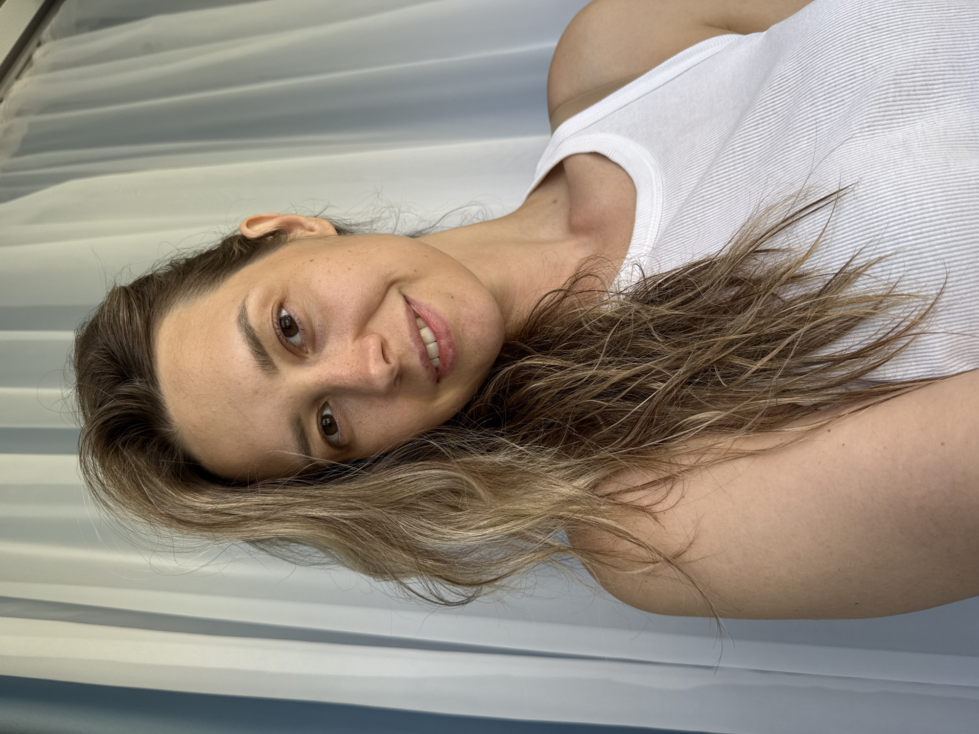 A woman with light brown hair is pictured from the chest up, looking directly at the camera and smiling. She is wearing a white ribbed tank top. The background consists of light-colored curtains with vertical pleats. The lighting appears to be natural, coming from the right side of the frame. The woman is positioned in the center of the image, with her hair falling over her left shoulder and across her chest.