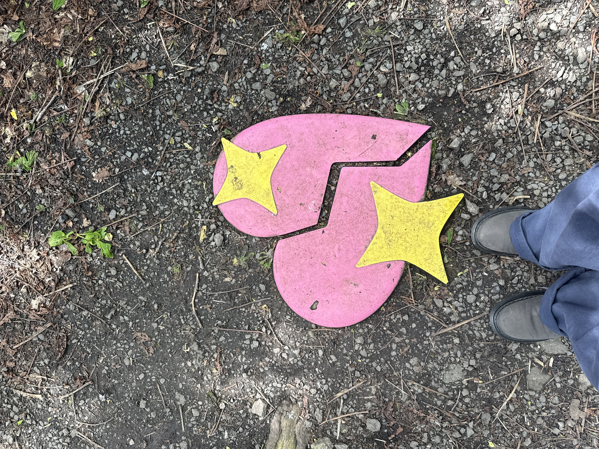 A high-angle, outdoor shot shows a broken, pink, heart-shaped object adorned with yellow stars on a ground covered in gravel and debris. The heart is split into two pieces with the yellow stars situated on each segment. The ground is composed of small pebbles, dirt, and scattered twigs and leaves. The lower right corner of the image features the feet and lower legs of a person wearing gray shoes and blue pants. The location appears to be in a natural or semi-natural area, such as a park or garden.