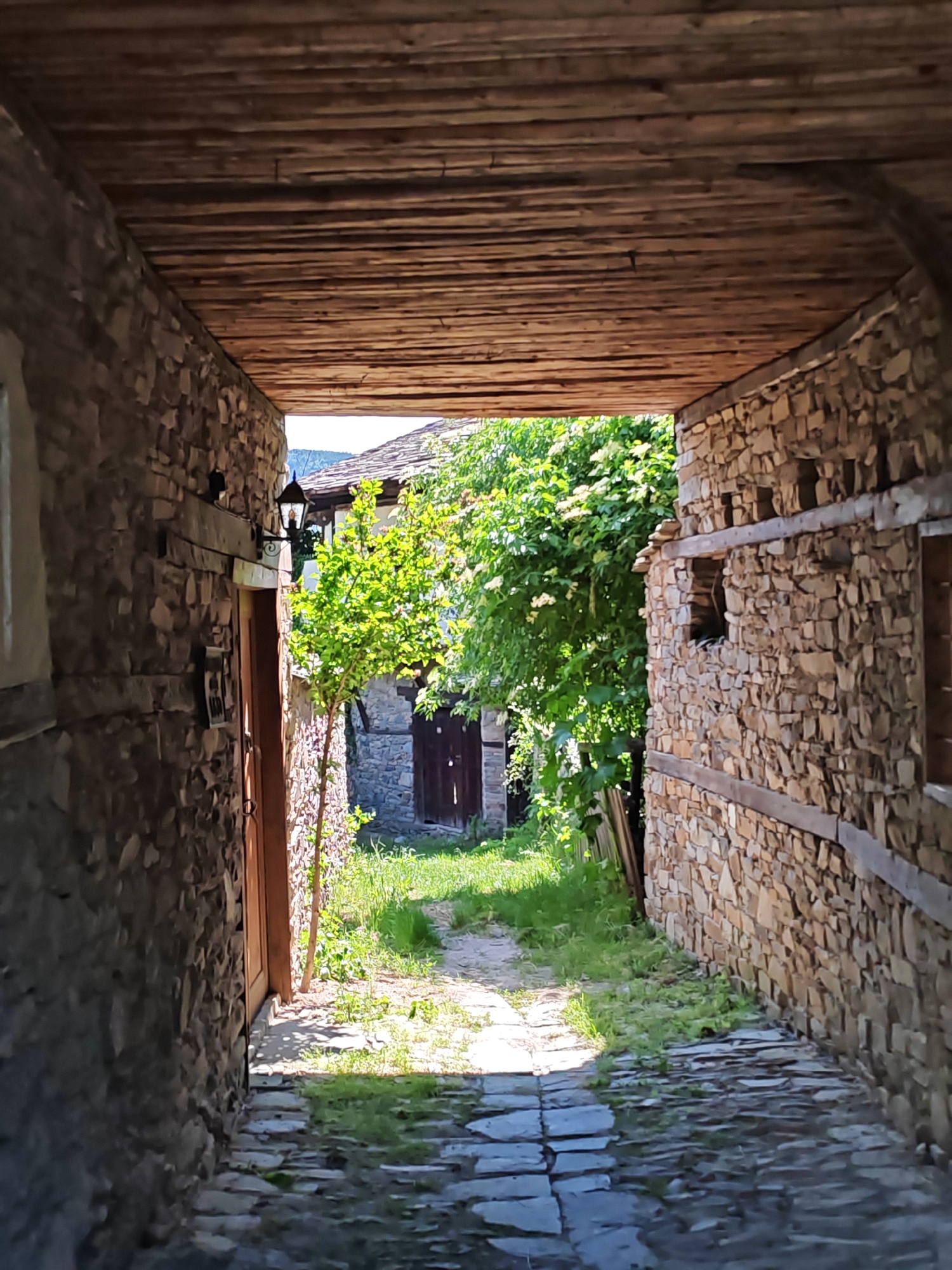 The image depicts a narrow, covered passage in the village of Kovachevitsa, Bulgaria, leading to an open, sunlit outdoor area. The passage is constructed with rough-hewn, light-colored stone walls on both sides and a ceiling composed of numerous horizontal, rough-cut wooden planks.
The left wall of the passage features a dark wooden door, partially obscured by shadow. Above and to the right of this door, a black, wrought-iron lantern with a glass enclosure is affixed to the stone wall. Further along the left wall, towards the foreground, an indistinct dark object is visible.
The right wall of the passage is also constructed from irregular stone and incorporates horizontal wooden beams. It contains a small, irregularly shaped opening or niche, and further towards the foreground, a dark, rectangular opening, possibly a window or another niche.
The floor of the passage is paved with flat, irregular stone slabs, which transition into a dirt path with patches of green grass at the exit.
Beyond the passage, the scene is brightly illuminated by natural light. A small, vibrant green-leaved tree stands immediately outside the left wall, casting a shadow. Further into the background, lush green foliage with small white flowers is visible on the right. Traditional Bulgarian architecture is evident in the background, with a stone-walled building featuring a dark wooden door and a stone-shingled roof. Part of another stone roof and distant, hazy hills under a clear sky can be seen in the upper background. No people or animals are present in the image.