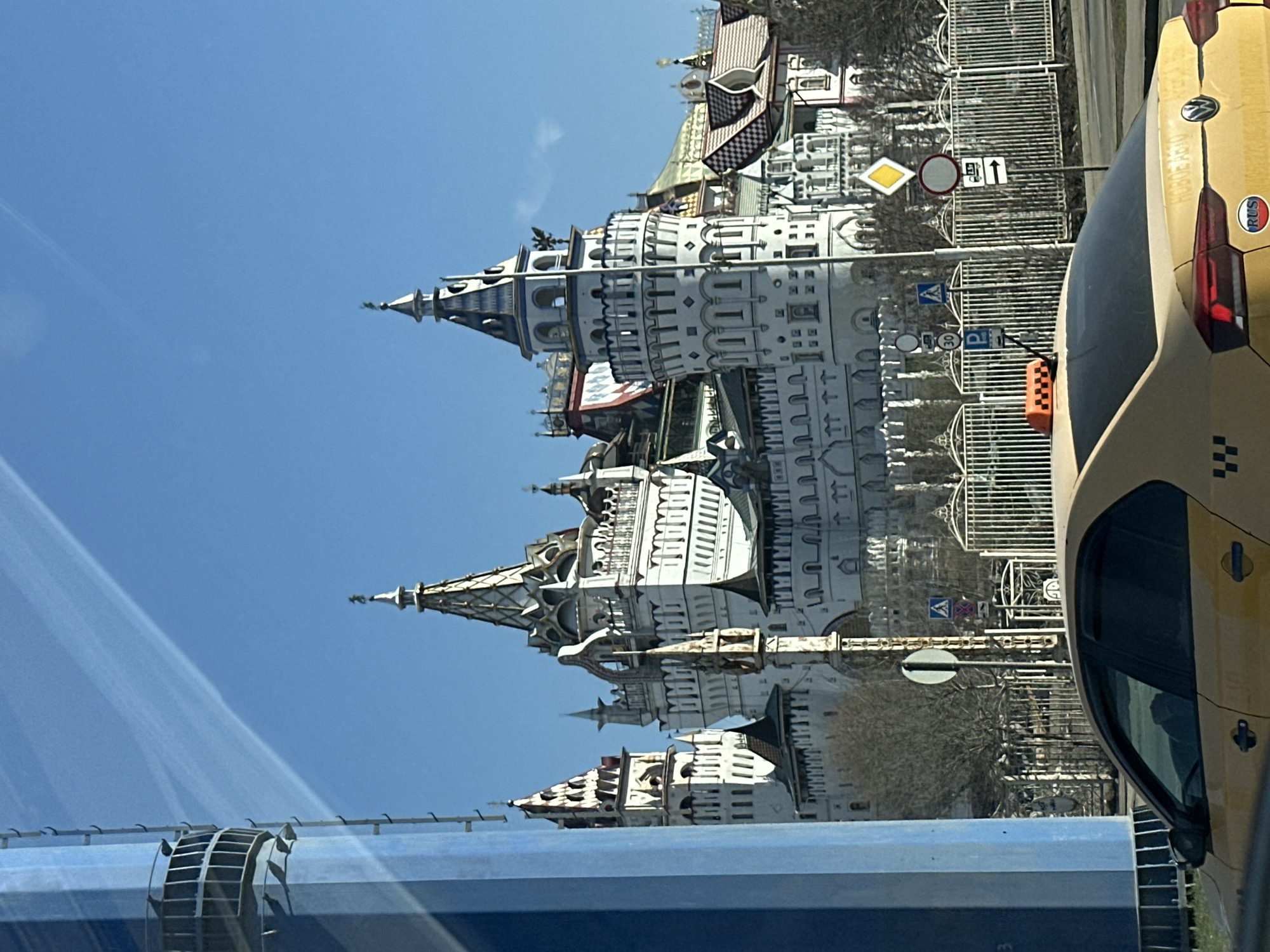 The image shows a view of the Izmailovo Kremlin in Moscow, Russia, through what appears to be the window of a vehicle, likely a bus or car. The Kremlin's distinctive white and ornate towers are visible against a clear blue sky. A yellow taxi cab is partially in view on the right side of the frame. A blue structure is visible on the lower left. Street signs and fencing can also be seen in the background.