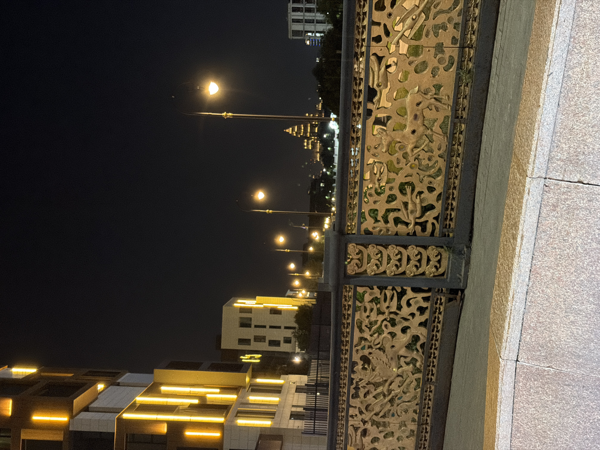 A nighttime view from a bridge in Astana, Kazakhstan, showcases a city skyline illuminated by streetlights and building lights. In the foreground, a decorative metal railing features intricate carvings of animals, possibly depicting scenes from Kazakh folklore. Beyond the railing, a walkway separates it from the street. The middle ground reveals several modern buildings, some with vertical yellow lights accentuating their facades. Further in the distance, a prominent building with a dome-like structure is brightly lit, and other buildings line the horizon. Streetlights with warm, glowing bulbs are visible along the street. The sky is dark, indicating it is nighttime. The overall scene conveys a sense of urban development and cultural representation through the architectural and decorative elements.