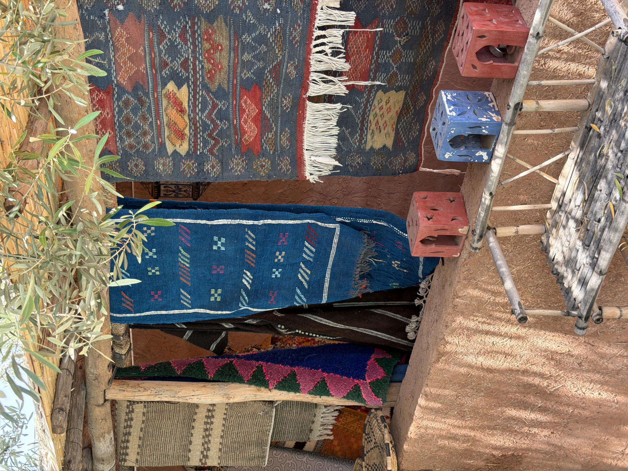 This vertically oriented image captures a vibrant display of traditional textiles and crafts in Tahannaout, Morocco. Numerous intricately patterned rugs and runners are prominently featured, hung from wooden beams. The uppermost rug is dark blue with elaborate geometric designs in red, brown, and cream, and has white fringed edges. Below it, a bright blue runner with smaller, colorful motifs hangs. Further down, various other textiles are visible, showcasing diverse patterns and colors including green, pink, black, and beige.
On the left side of the frame, a thriving olive tree with slender green leaves and wooden branches partially obscures the view, adding a natural element to the scene. To the right, three cubic, perforated stools are arranged next to a bamboo ladder or scaffolding. Two stools are red and one is blue, all appearing weathered with decorative cut-outs. The ground or a wall on the right is a rough, reddish-brown earthen surface, likely made of rammed earth or mudbrick, with visible straw fibers.
The scene is bathed in bright sunlight, indicated by the clear illumination and the shadows cast by the bamboo ladder on the earthen surface, suggesting a sunny daytime. The overall impression is one of a lively, traditional outdoor setting, possibly a market or workshop, highlighting local Moroccan craftsmanship and natural materials. No visible text is present in the image.
