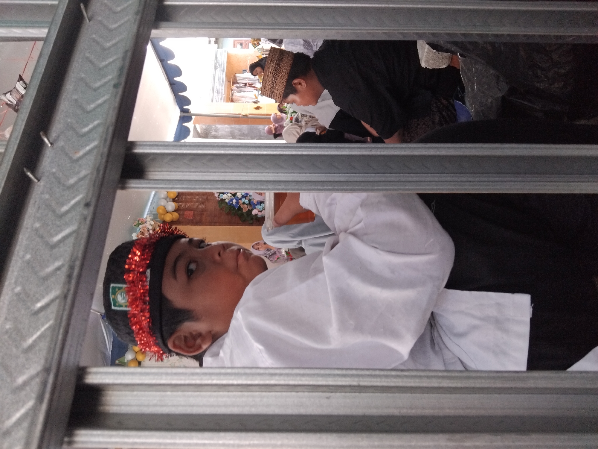 A young boy with dark hair wears a white collared shirt and black pants, adorned with a red garland on his head. He is positioned behind a metal gate, looking to his right with a neutral expression. In the background, other individuals are visible, some wearing headscarves. The scene appears to be indoors, possibly a community gathering, with decorations such as balloons visible in the distance. The environment suggests a religious or cultural event. The overall lighting is dim, with a slight glare on the metal bars.