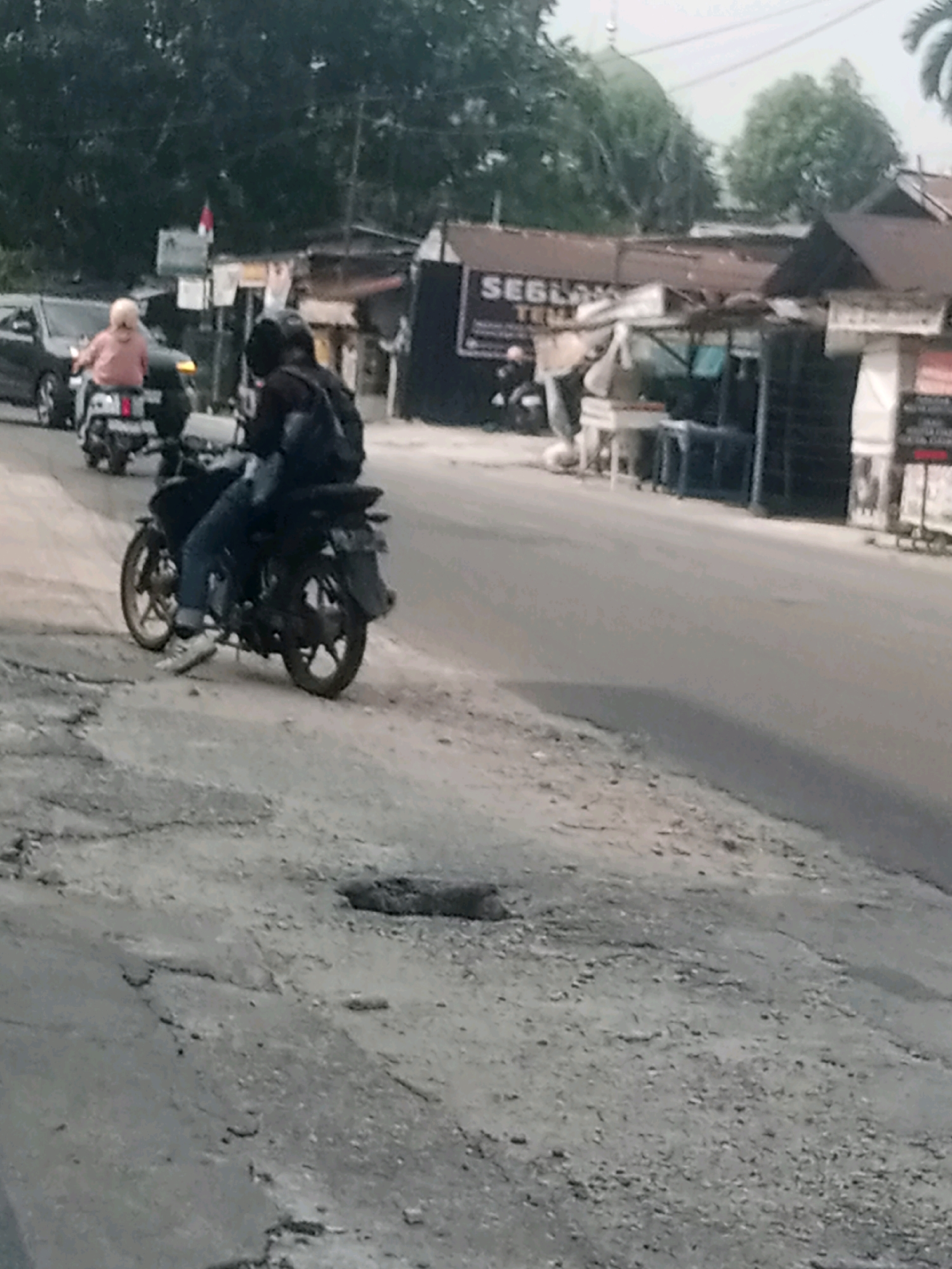 The image depicts a street scene in Pekanbaru, Indonesia, likely during daylight hours with overcast conditions. In the foreground, a motorcycle with two occupants is stopped or moving slowly on a rough, uneven road surface, characterized by cracks and small stones. The driver wears a helmet and blue jeans, with white sneakers. The passenger, positioned behind the driver, also wears a helmet and a dark backpack. To the left of the motorcycle, another motorcycle is visible, carrying a person wearing a pink shirt and a headscarf. A dark-colored car is partially visible behind this second motorcycle. In the background, several roadside structures with corrugated metal roofs suggest a commercial or informal settlement area. A sign with "SEBLONK" written on it is discernible on one of these structures. Trees line the background, and a dome, possibly part of a mosque, is visible in the upper portion of the image, partially obscured by foliage. The overall impression is a snapshot of everyday life on a street in Pekanbaru.