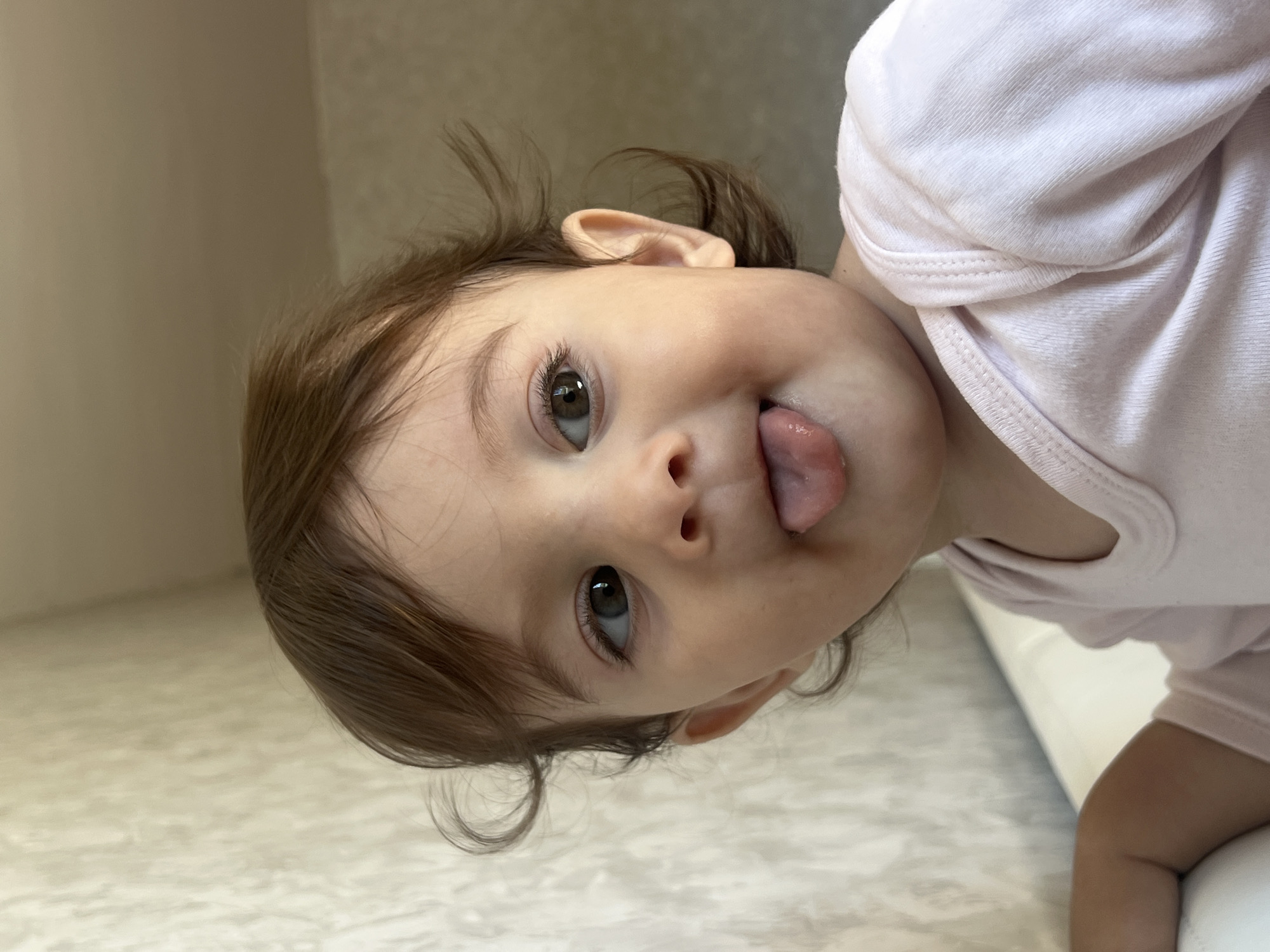 A baby with brown hair and brown eyes is sticking out their tongue. The baby is wearing a light pink bodysuit. The baby is lying on a white surface, possibly a sofa. The location is unknown and could not be determined to be Elena, Bulgaria.