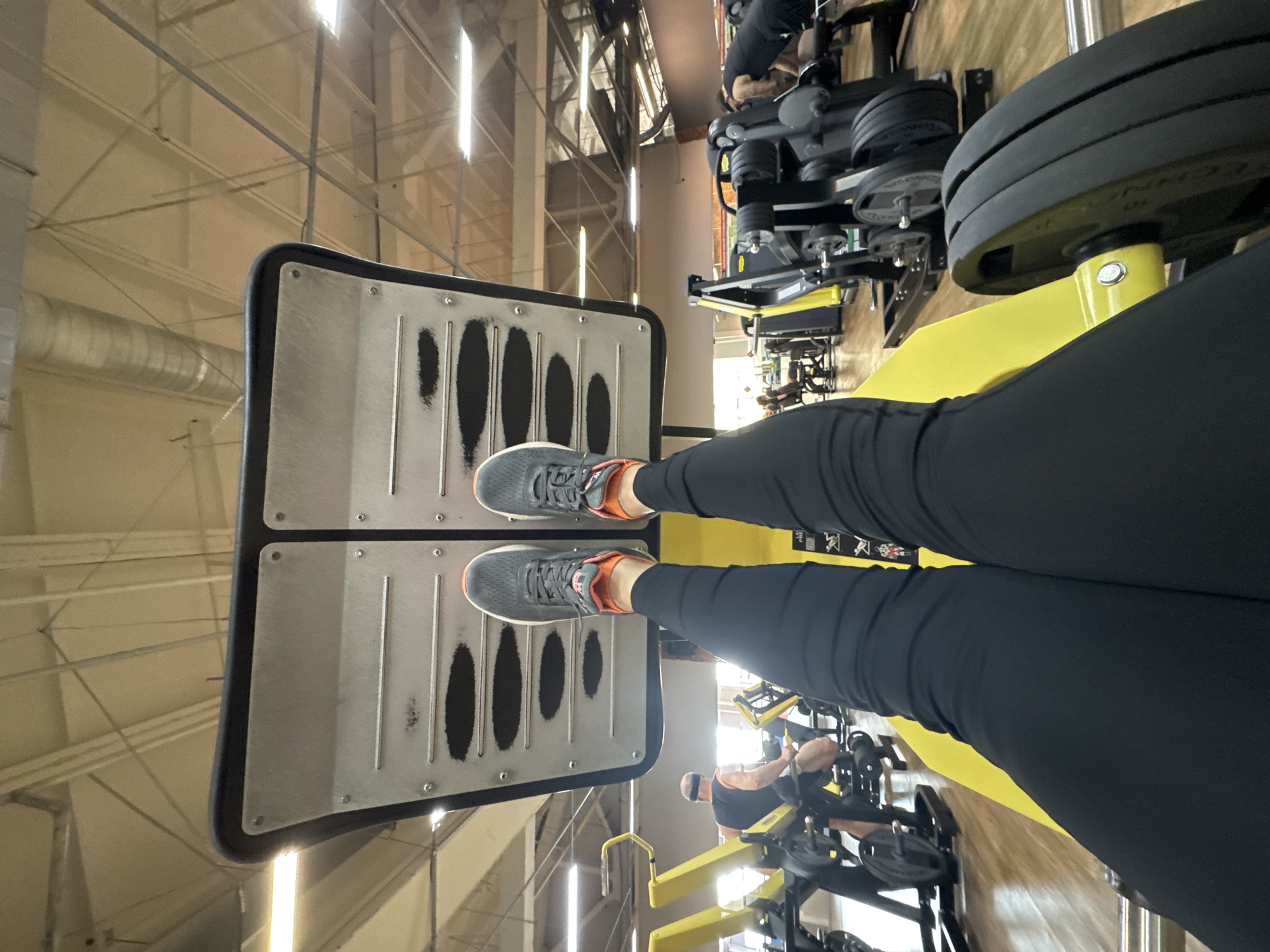 A person wearing black leggings and gray and orange athletic shoes is positioned on a leg press machine in a gym. The machine has a silver footplate with black oval cutouts and is attached to a yellow frame with black weight plates. The gym's interior features a metal ceiling structure and various other exercise machines, some of which are occupied by other gym patrons. The location is the city of Chaikovsky, Russia.