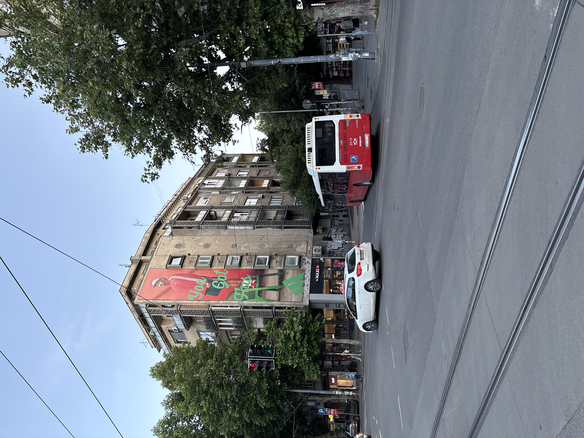 This is a photograph taken from a street level perspective in Belgrade, Serbia, capturing a bustling intersection with urban elements. The foreground shows tram tracks embedded in asphalt, with a grey road surface. A white station wagon is captured in motion, navigating the intersection. To the right, a red and white public bus is also in motion, its rear visible. The background is dominated by a multi-story apartment building with a large advertisement plastered on its facade, depicting a man in a red tracksuit. Adjacent to the building are trees providing shade. A traffic light, displaying red and green signals, stands to the left of the frame. Several pedestrians are visible on the sidewalks, and other vehicles are present in the distance, suggesting a typical urban scene. The sky above is clear and a light blue, indicating it's likely late afternoon or early evening.