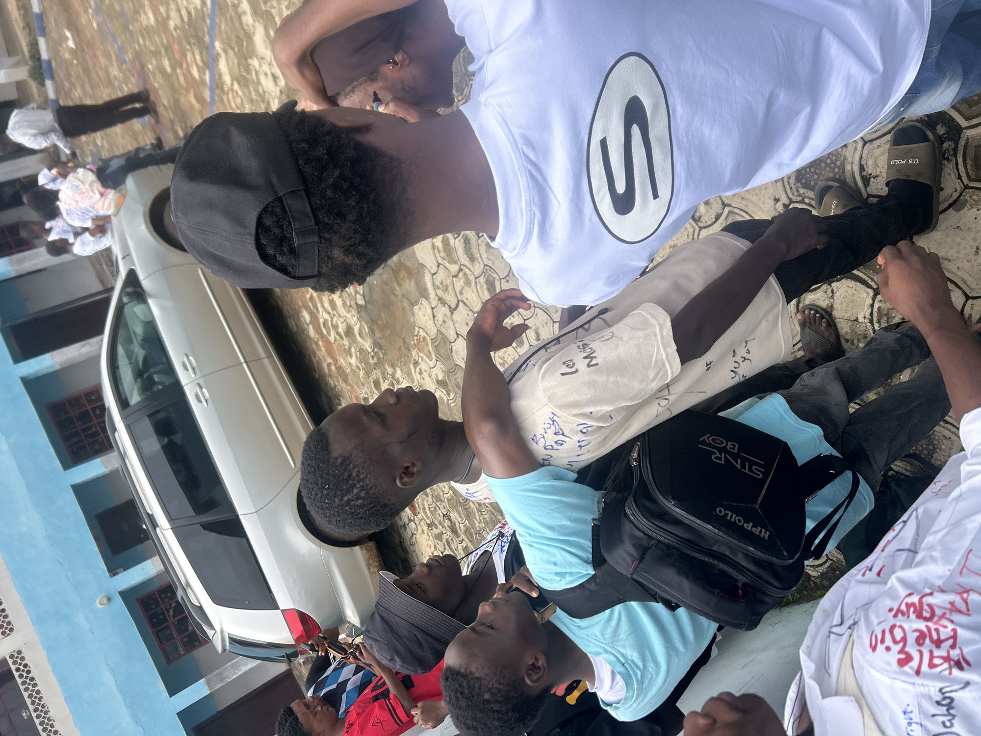 The image captures a group of young Black men gathered outdoors, likely in Ikorodu, Nigeria. The scene is casual and appears to be during daylight hours, with the ground appearing wet, suggesting recent rain or a damp environment.
In the foreground, several individuals are visible, some wearing t-shirts and casual attire. One person in the center wears a white t-shirt with a prominent black "S" logo enclosed in a circle. Another person to their right is wearing a light blue t-shirt and has a black backpack with "STAR BOY HPPOILO" written on it. Further to the left, a person in a black baseball cap and a white t-shirt is seen. Other individuals are partially visible, some with messages written on their clothing, such as "the bin guy" and "Jahon" in red marker. A white car is parked on the left side of the frame, with a blue building in the background. The ground is paved with interlocking bricks, some of which are wet. The overall mood seems relaxed and social, with the individuals appearing to be interacting or posing for the photo.