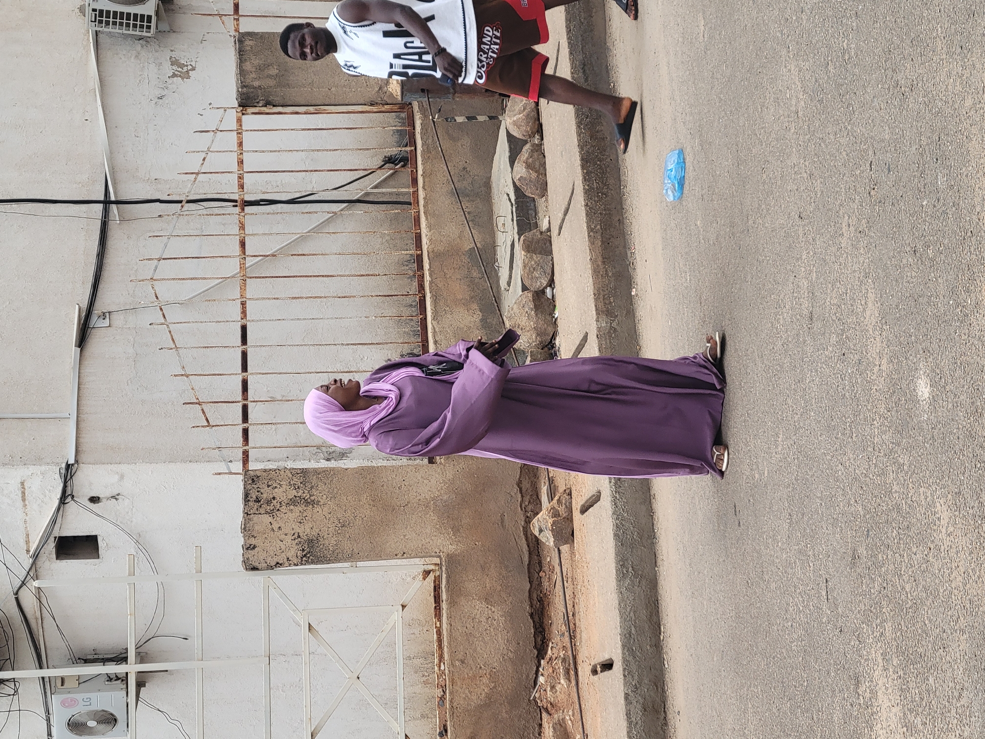 The image shows a street scene in Jos, Nigeria. A woman wearing a long purple dress and head covering is walking along the right side of the road. A man in a white basketball jersey and red shorts stands near a rusty metal fence. An air conditioning unit is visible on the side of a white building. A blue plastic bag is visible on the road.