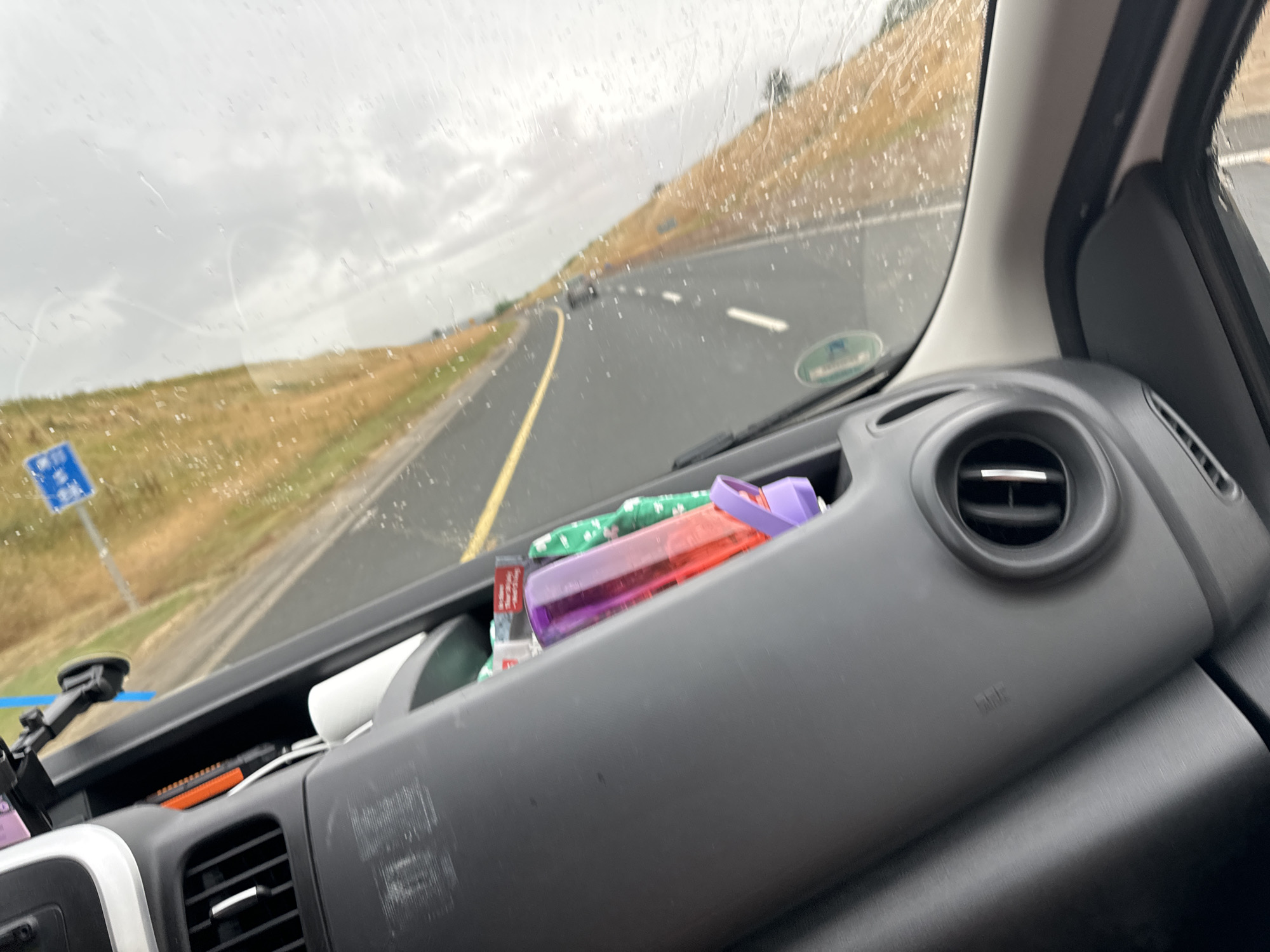 The image depicts the interior of a vehicle, with a focus on the dashboard and the view through the windshield. The dashboard is black and grey, featuring an air vent and a textured surface. On the dashboard, a red and purple bottle is visible, along with a roll of white material and a bright orange object. The windshield shows a road with a yellow line, indicating a highway, and sparse traffic in the distance. The weather appears overcast with rain streaks on the windshield. The landscape outside is characterized by dry grass and a cloudy sky. There is a blue road sign with white symbols visible in the distance. Based on the context of Tubberneering, Ireland, this scene is likely captured while driving on a road in or around that location.