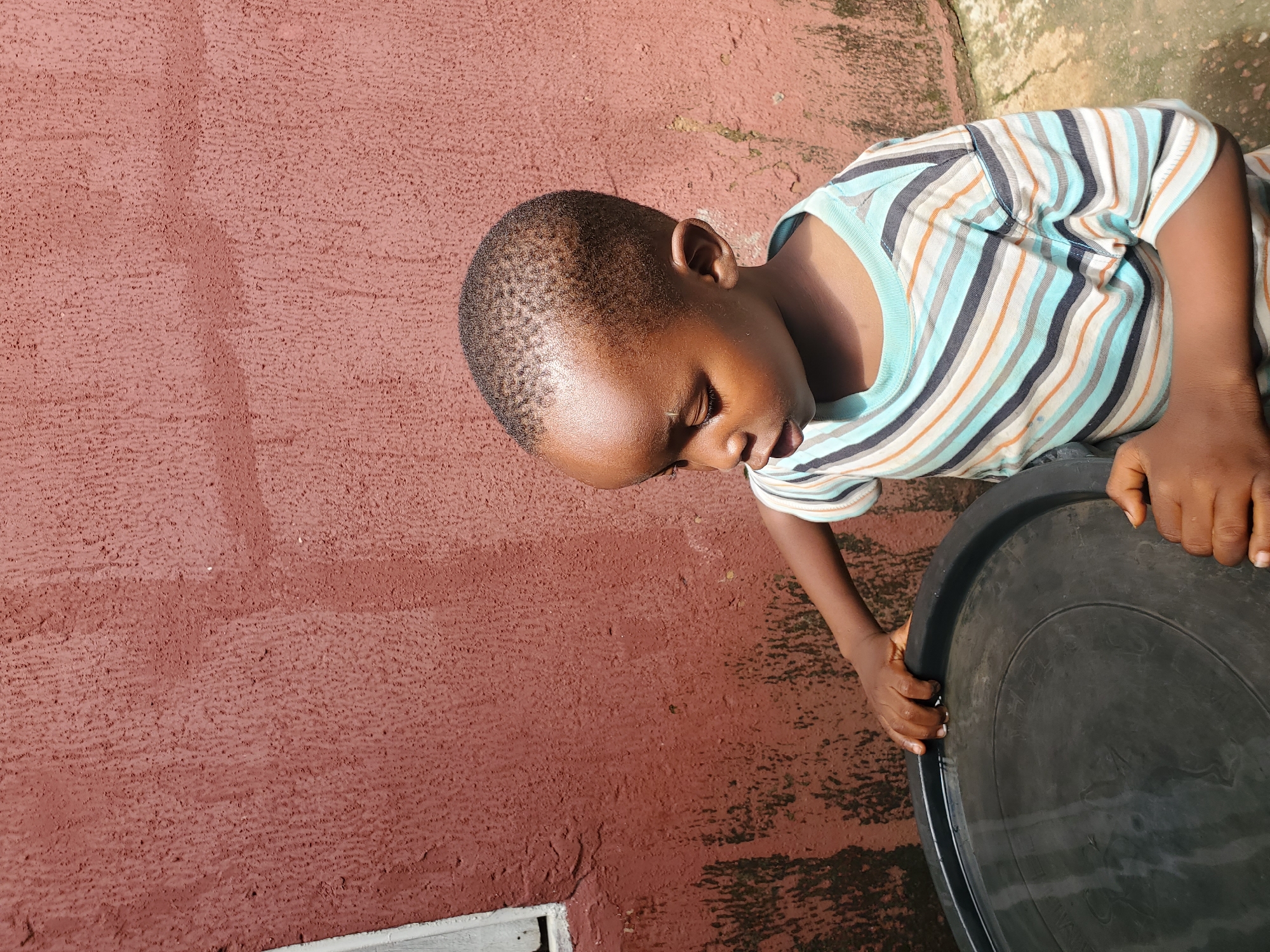 A young boy, with dark skin and short, cropped hair, is positioned in the foreground, looking down and to his left. He is wearing a horizontally striped t-shirt in shades of blue, white, gray, and orange. His hands are placed on the rim of a large, black, circular object, possibly a container or basin, which contains a small amount of water. The boy's gaze is directed towards this object.
The background features a textured, reddish-brown wall, with some areas of discoloration and what appears to be moss or algae growth. To the left of the boy, a sliver of a light-colored window frame is visible. The ground beneath the boy is a mix of concrete and dirt. The scene is brightly lit, suggesting it is daytime. The environment appears to be outdoors, likely in a residential or communal outdoor space. The scene is devoid of any clear indicators placing it specifically within the city of Jos, Nigeria, beyond what might be inferred from the general architectural style or environmental conditions.
