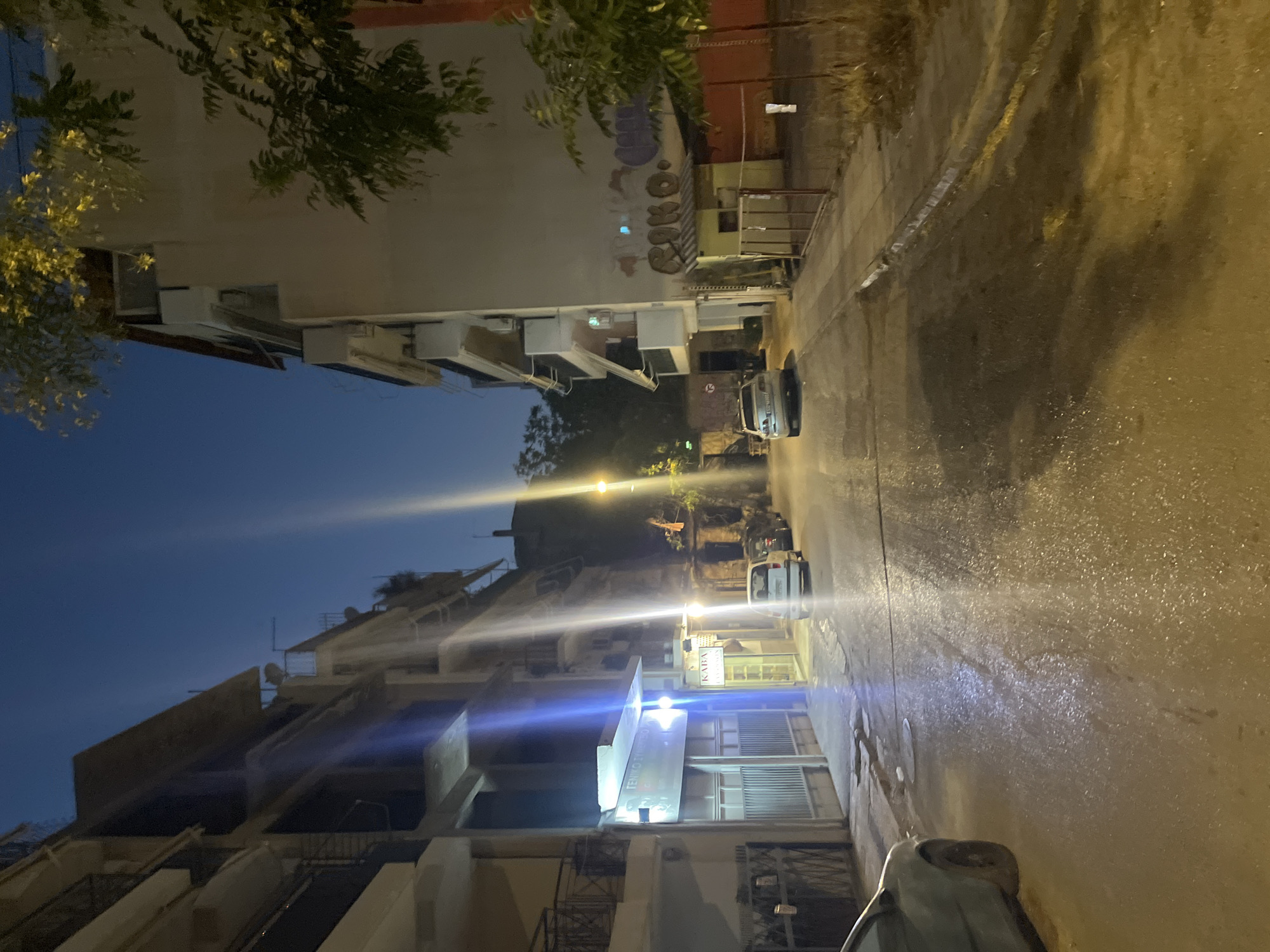 This image depicts a street scene in Athens, Greece, during the late evening or early night. The street is wet, reflecting the light from street lamps and buildings, suggesting recent rain or street cleaning. Buildings line both sides of the narrow street, appearing to be residential or commercial structures with multiple stories. Some buildings feature balconies, while others have storefronts or entrances at street level.
On the left side of the street, there's a building with a large metal gate in the foreground. Further down, a business with a sign that reads "ΓΕΝΙΚΟ ΣΥΜΒΟΥΛΙΟ" is visible, with bright lighting illuminating the facade. Adjacent to it, another storefront has a sign that appears to read "KABA". A white van is parked on the left side of the street, partially obscured by the building.
On the right side of the street, a larger building dominates the scene, with graffiti visible on its beige facade, including the word "RINO" and other abstract markings. A small, yellow, kiosk-like structure with a pitched roof is situated in front of this building, behind a metal fence. Further down the street, a few cars are parked along the curb, including a silver sedan and a white SUV.
The sky is a deep blue, transitioning to a lighter hue near the horizon, indicating the twilight period. Trees with yellowing leaves are visible at the top of the image, near the buildings on the right. The overall atmosphere is calm and quiet, with no people visible in the frame. The scene suggests a typical urban environment in Athens, illuminated by artificial lights.