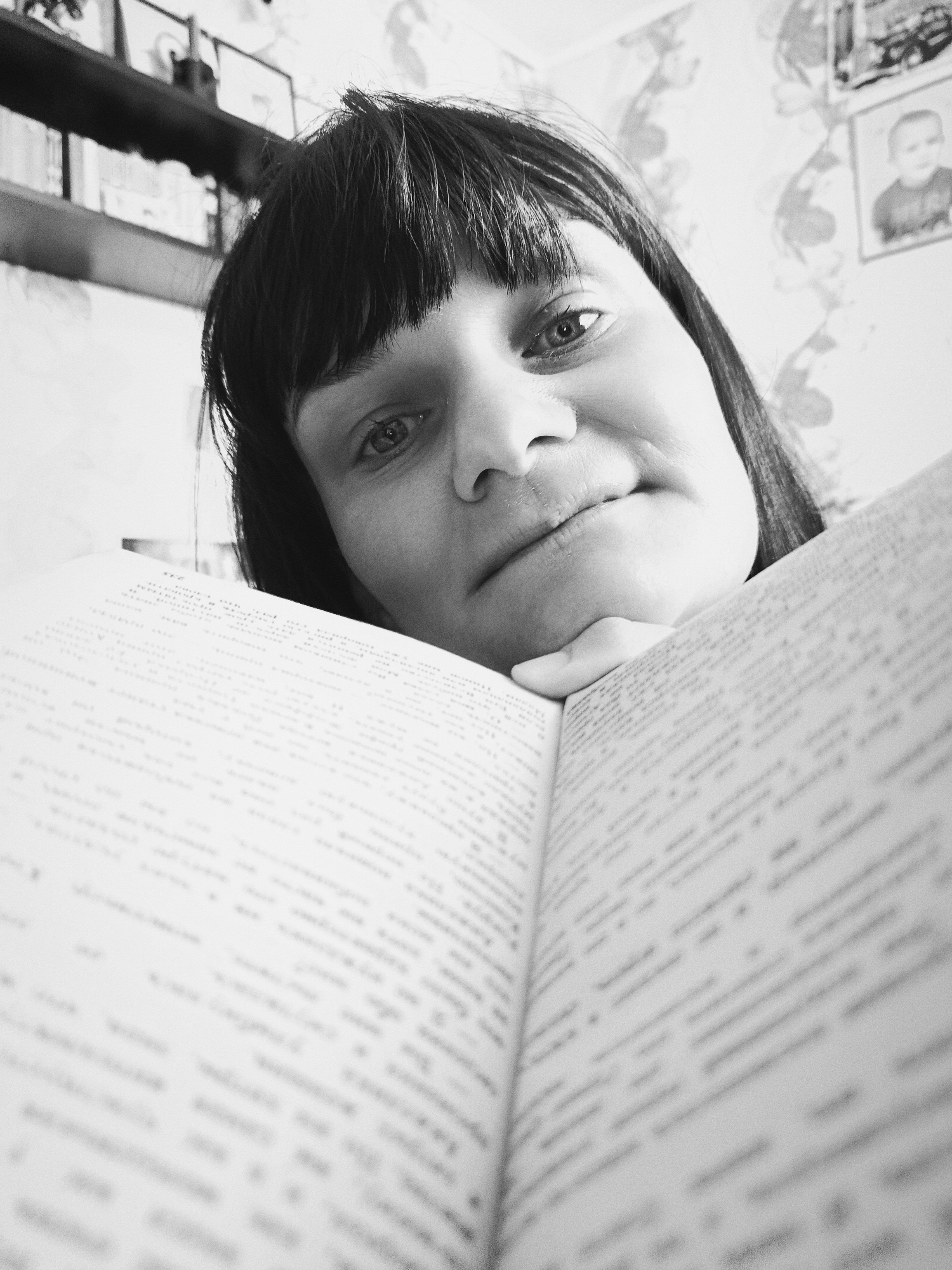 A black and white, close-up, eye-level shot depicts a woman with dark hair and bangs, holding an open book. Her face is angled downwards towards the book, resting her chin on the edge of the pages. The book takes up the majority of the foreground, with blurred text visible on the pages. Behind the woman, a shelf with framed pictures is partially visible, along with floral patterned wallpaper. The location in the city is Unknown, Unknown.