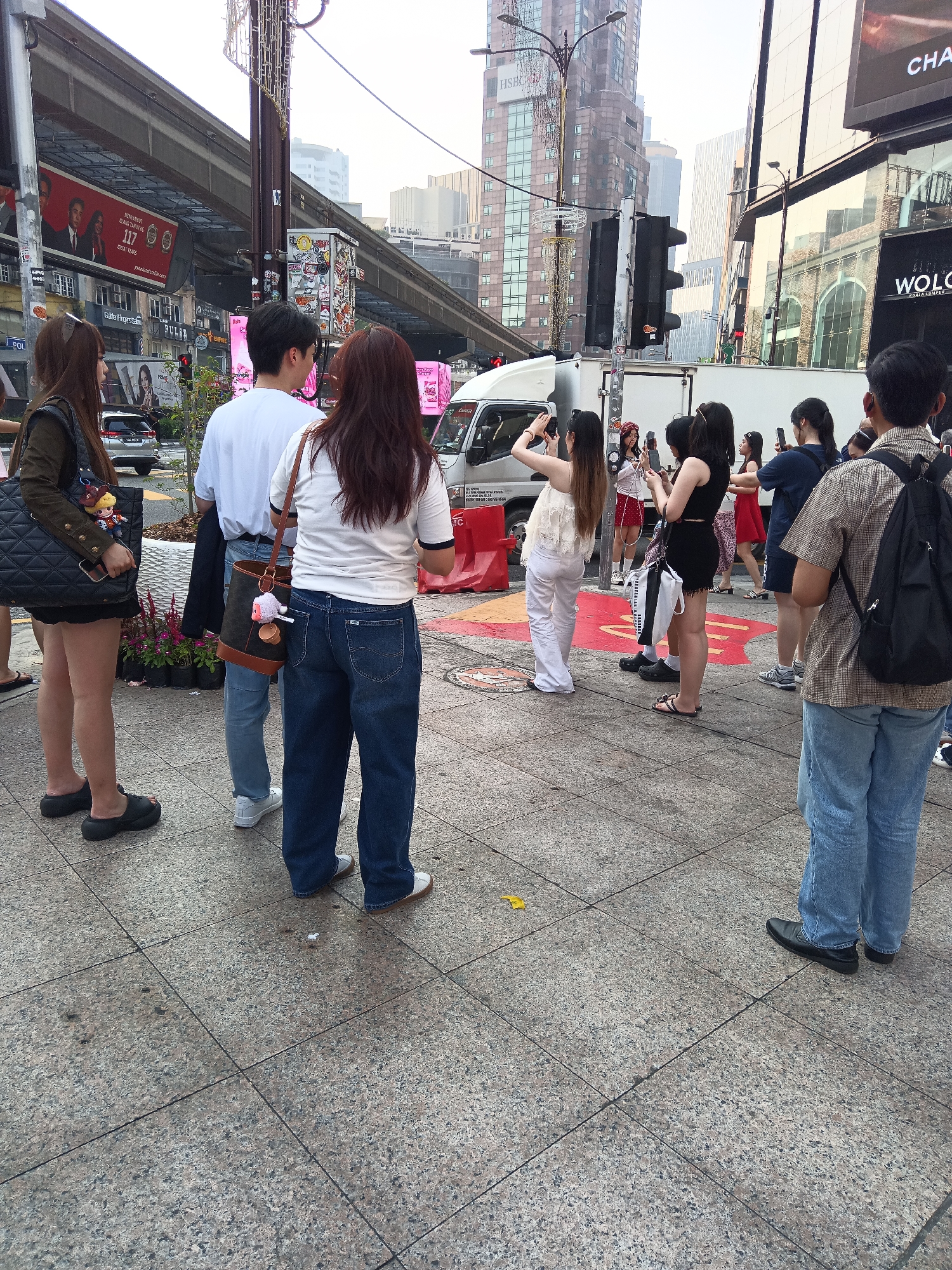In Kuala Lumpur, Malaysia, a group of people are gathered on a sidewalk near a street. The scene is characterized by urban elements, including an elevated train track, tall buildings, traffic lights, and street advertisements.
Several individuals are visible, with their backs primarily turned towards the viewer. Some are holding mobile phones, suggesting they might be taking pictures or recording videos. One woman in the foreground is wearing a white t-shirt and blue jeans, carrying a brown bag with a light-colored charm. To her left, a man in a white t-shirt and blue jeans also has a bag with a charm. On the far left, a woman in a dark jacket and shorts carries a large black tote bag.
In the background, a white truck is parked, and other people are positioned further down the sidewalk, some also engaged with their phones. The environment features a mix of concrete pavement with visible grid lines, potted plants, and various commercial signage on the buildings. The sky appears overcast.