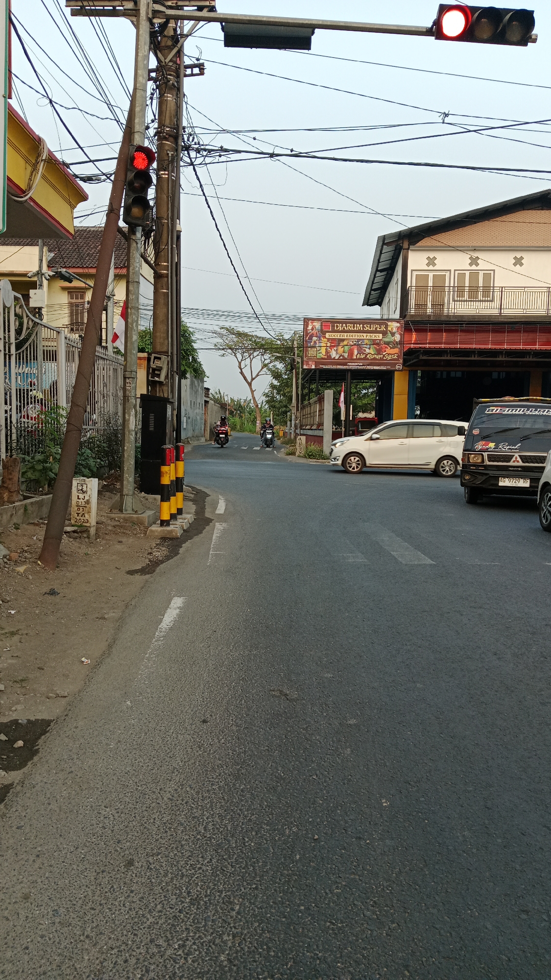 The image depicts a street scene in Blitar, Indonesia, with a traffic light showing a red signal. The traffic light is mounted on a utility pole, which is adorned with numerous electrical wires. To the left of the pole is a building with a yellow facade and a metal fence. On the right side of the road, there's a white car and a black van with a license plate AG 9729 RF. Further down the road, two motorcycles are visible, each with a rider. A billboard advertising "Djarum Super Soccer Edition Packs" is prominent on the right side. The sky is overcast, suggesting it is either early morning or late afternoon. The road is paved and appears to be a two-lane street. A pedestrian crossing is marked on the road. A small concrete marker with text is visible near the utility pole.