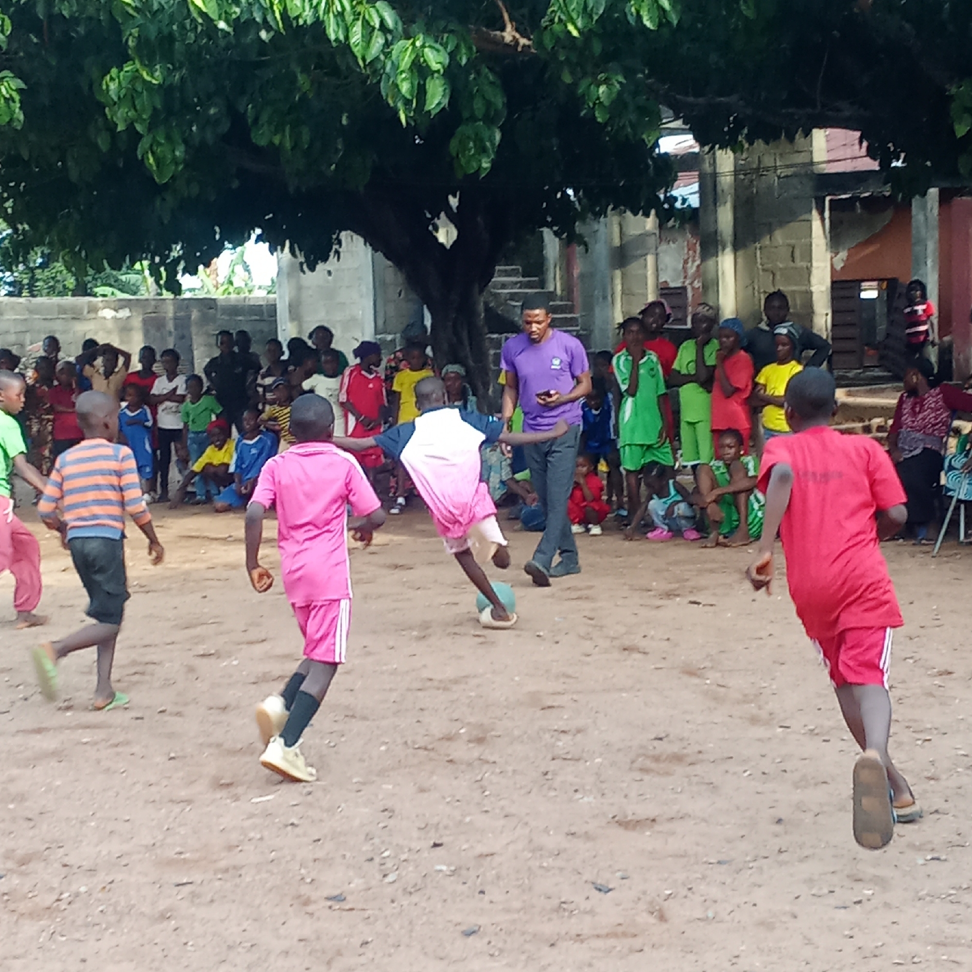 The image depicts a soccer game in Akwanga, Nigeria. Several children are playing on a dirt field under a large, leafy tree. A crowd of children and adults are watching the game. A man in a purple shirt appears to be supervising the game. A building stands in the background. The children are wearing different colored jerseys and shorts. Some are barefoot, while others wear shoes or sandals. The field is dry and dusty.