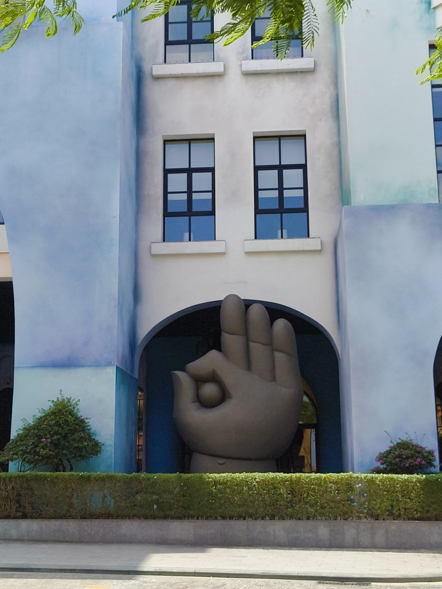 The image shows the exterior of a building with a large sculpture of a hand in the foreground. The building has a light blue and white facade with several windows. The hand sculpture, which is dark gray, is positioned under an archway. In front of the building, there is a neatly trimmed hedge and a paved area, possibly a sidewalk or road. Lush green foliage hangs from the top of the frame. The scene is located in Phú Quốc, Vietnam, and appears to be an outdoor public space or a resort. There are no people visible in the image.