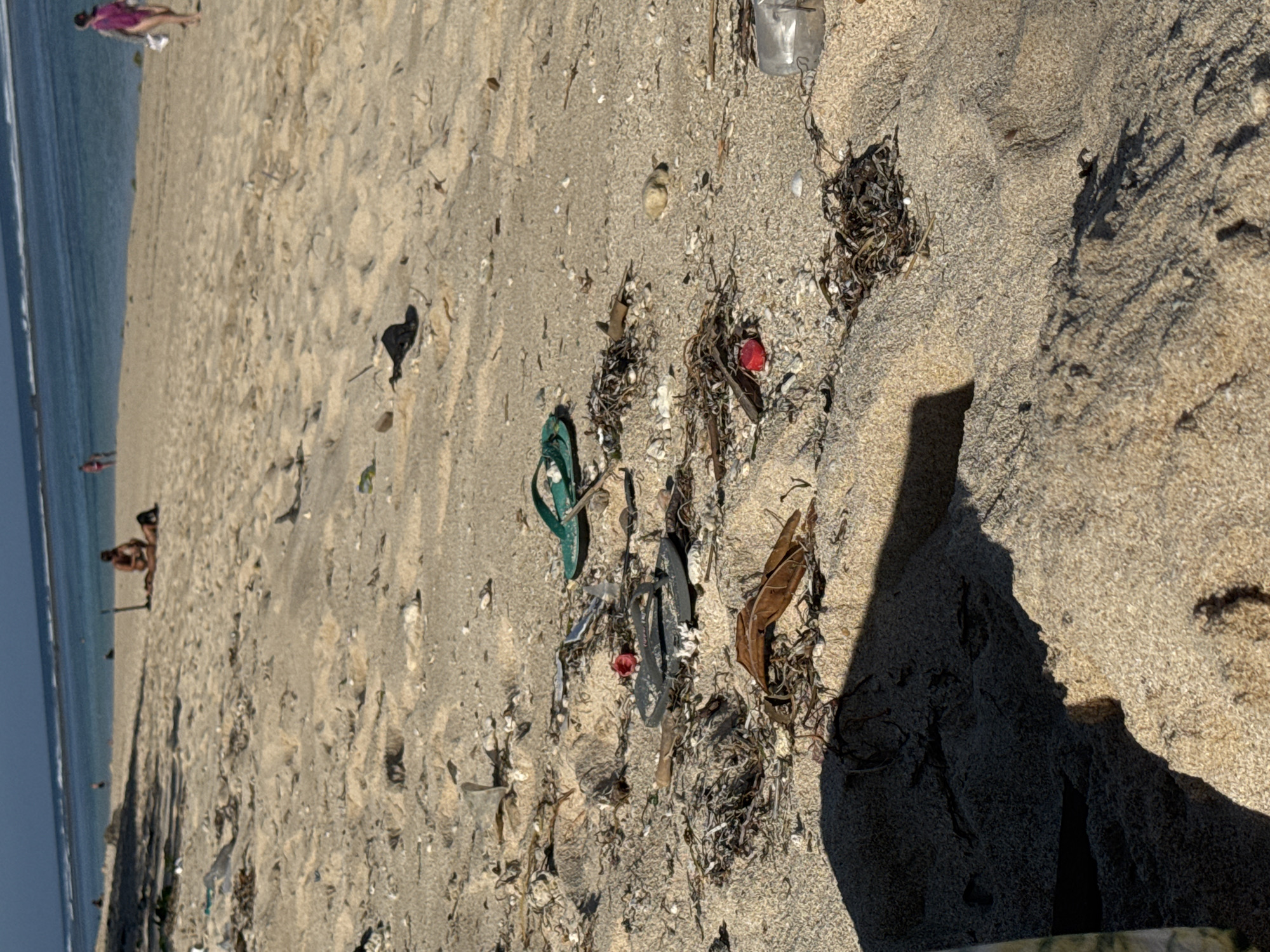This is a medium-angle, eye-level shot of a sandy beach in Kuta Selatan, Indonesia, with the ocean in the background. The beach is littered with debris including plastic bottles, seaweed, and flip-flops. In the distance, several people can be seen on the beach and in the water. One person, wearing a pink garment, walks along the shore, while a couple sits on the sand. The sand is light brown and textured, with some areas appearing darker due to shadows. The ocean is a deep blue, with small waves breaking near the shore. The sky is a clear, pale blue.