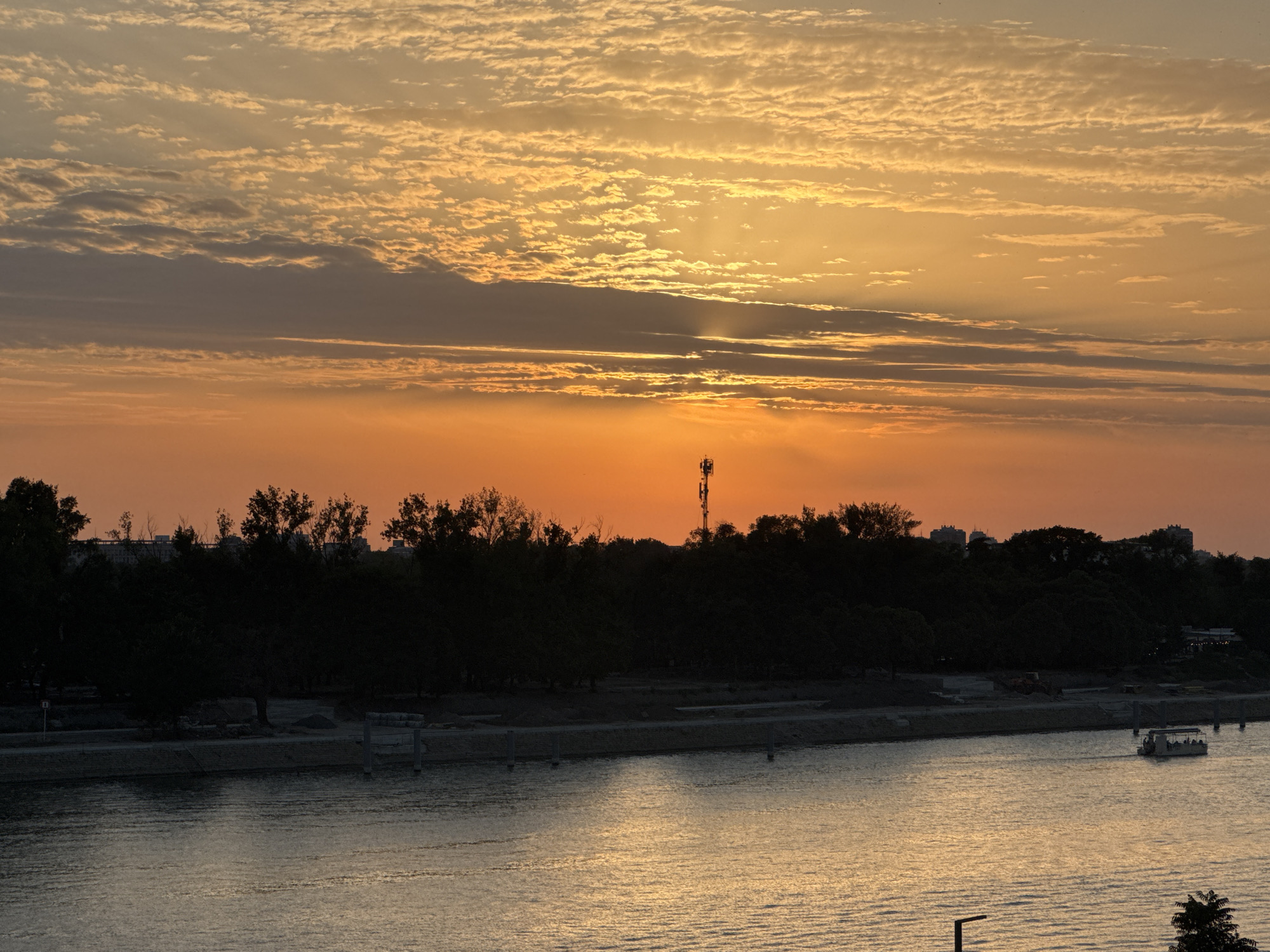 The image depicts a sunset over the Sava River in Belgrade, Serbia. The sky is filled with streaks of orange and yellow clouds, with rays of sunlight breaking through. A dark silhouette of trees and buildings lines the riverbank, punctuated by a tall cell tower. A small boat with several passengers is visible on the water, its reflection shimmering on the surface. The scene captures the serene beauty of a sunset over a bustling urban landscape.