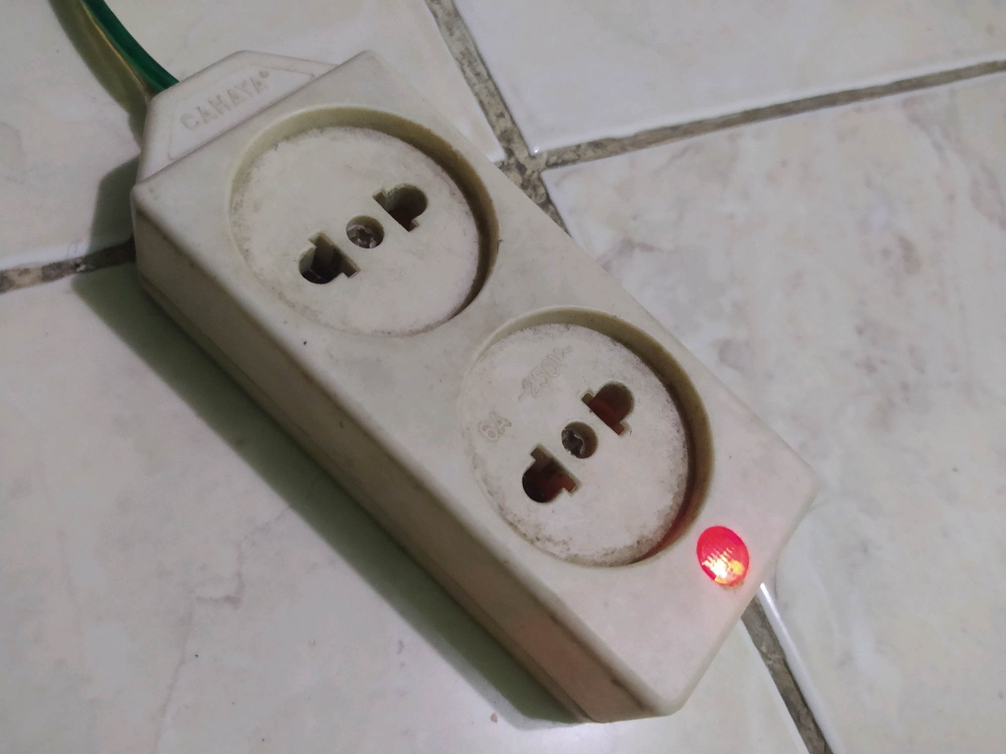 A close-up, eye-level shot focuses on a cream-colored, dual-outlet power strip positioned on a light-colored, tiled surface. The power strip is oriented diagonally from the upper left to the lower right of the frame. It bears the embossed brand name "CAMAYA" near the top. A green and yellow wire emerges from the left side of the power strip. Both outlets appear to be of the universal type, capable of accepting various plug configurations. One outlet is slightly obscured by shadow, while the other shows wear and dust accumulation. Below the right outlet, "6A - 250V~" is inscribed. A small, red indicator light on the right side of the power strip is illuminated, suggesting it is receiving power. The tiled floor exhibits grout lines and slight discoloration, typical of a domestic or commercial setting. The lighting is somewhat dim, casting a soft shadow from the power strip onto the tiled surface. The image is taken in Sroyo, Indonesia.
